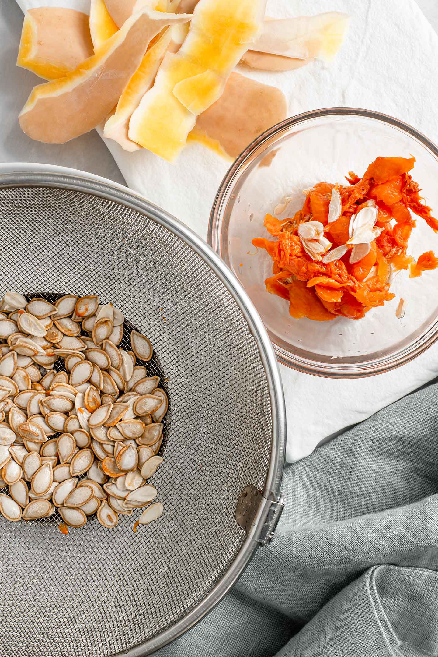 Butternut squash seeds are in a sieve with the stringy membrane in a separate small bowl. Slices of squash peel lay next to the seeds on a white tray.