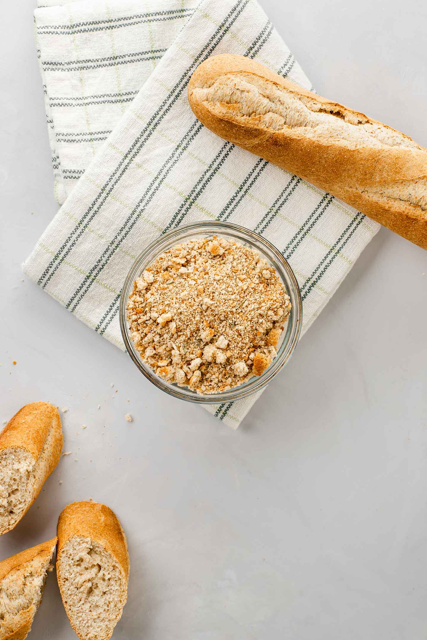 Top down view of quick and easy homemade breadcrumbs in a small glass bowl sitting on a dish towel with a baguette and slices of bread to the side