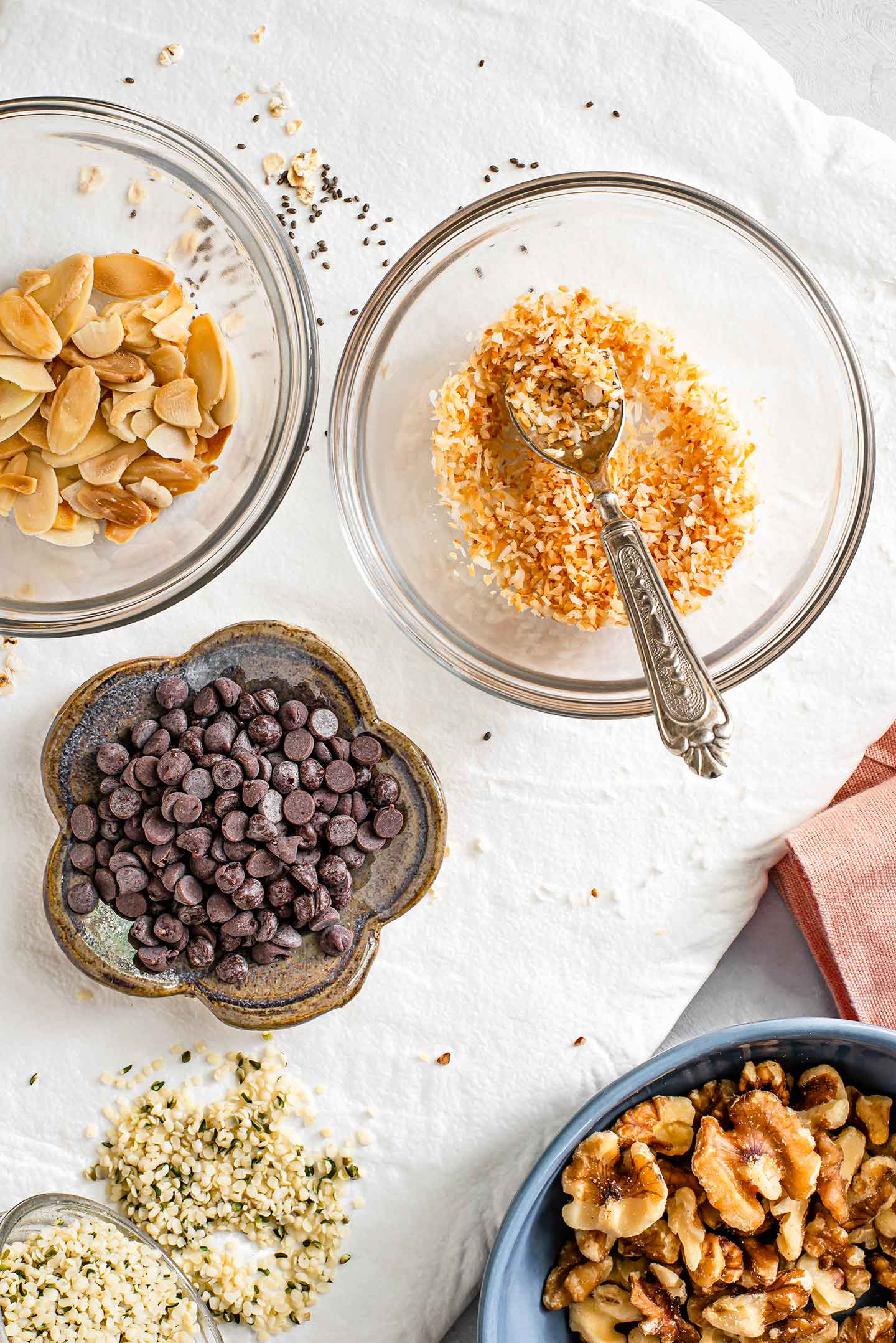 Top down view of toppings for overnight oats. Toasted almonds and coconut, chocolate chips, hemp seeds, and walnuts are pictured in separate dishes.