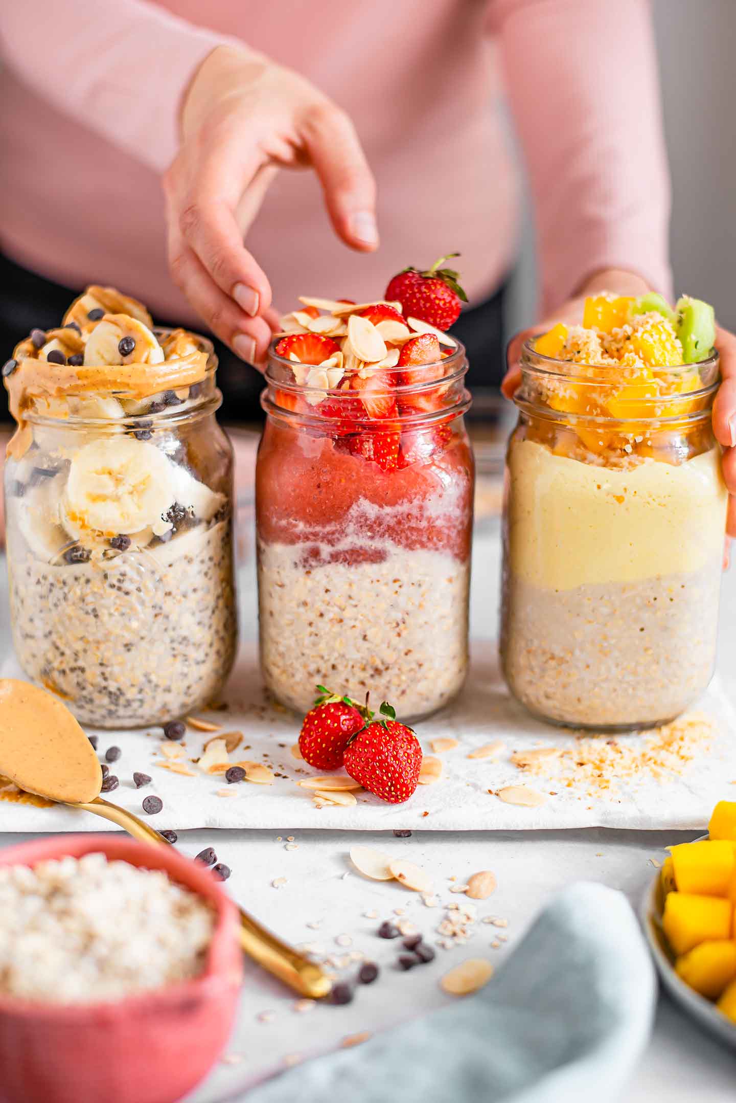 Side view of hands placing three jars of overnight oats on a white tray. One jar is topped with banana, chocolate chips, and runny peanut butter. The next jar is topped with strawberry jam, fresh berries, and toasted almonds. The final jar is topped with mango yogurt, fresh mango, and toasted coconut.
