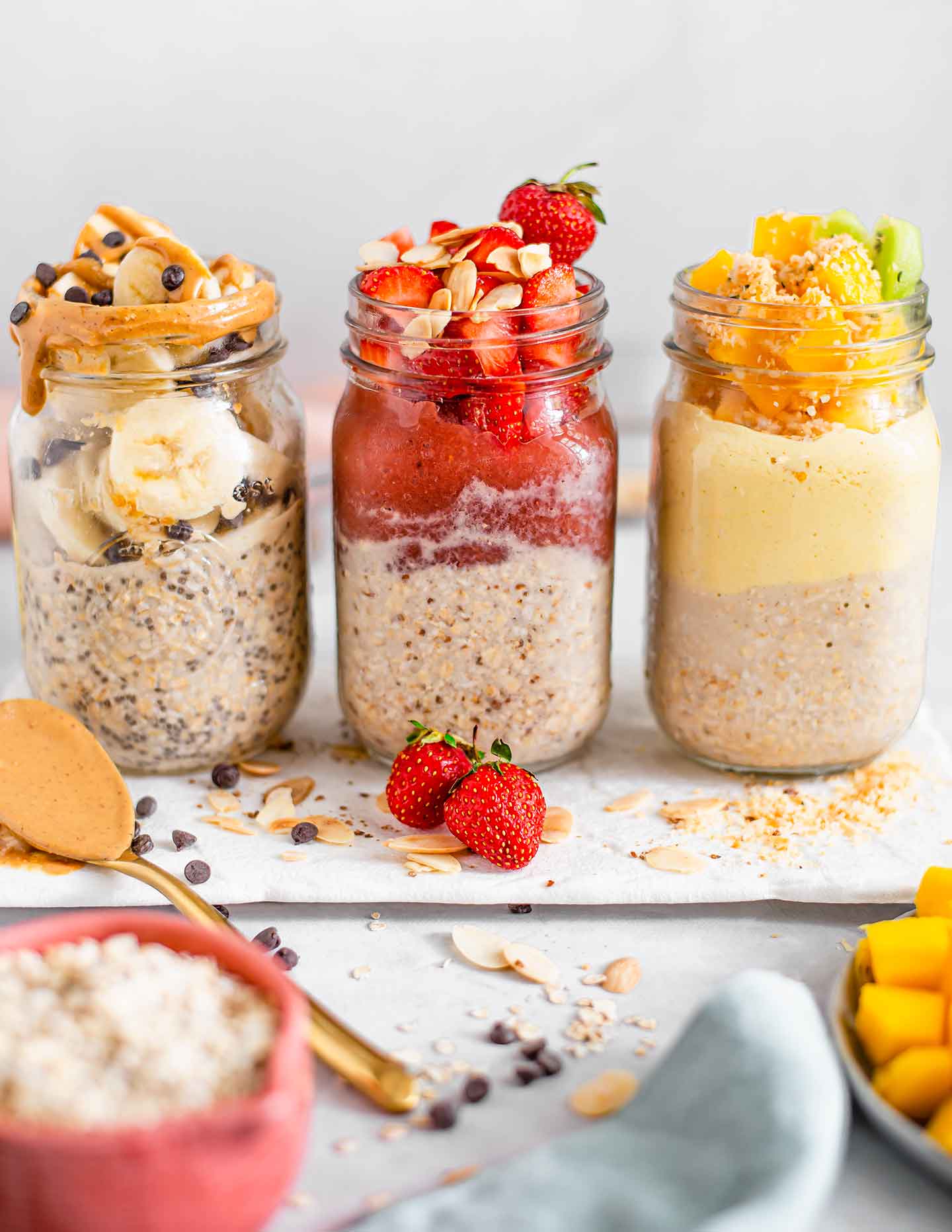 Side view of three jars of overnight oats. One is peanut butter banana chip, one is strawberries and jam, and the last is mango and yogurt. Ingredients are scattered around the base of the jars.