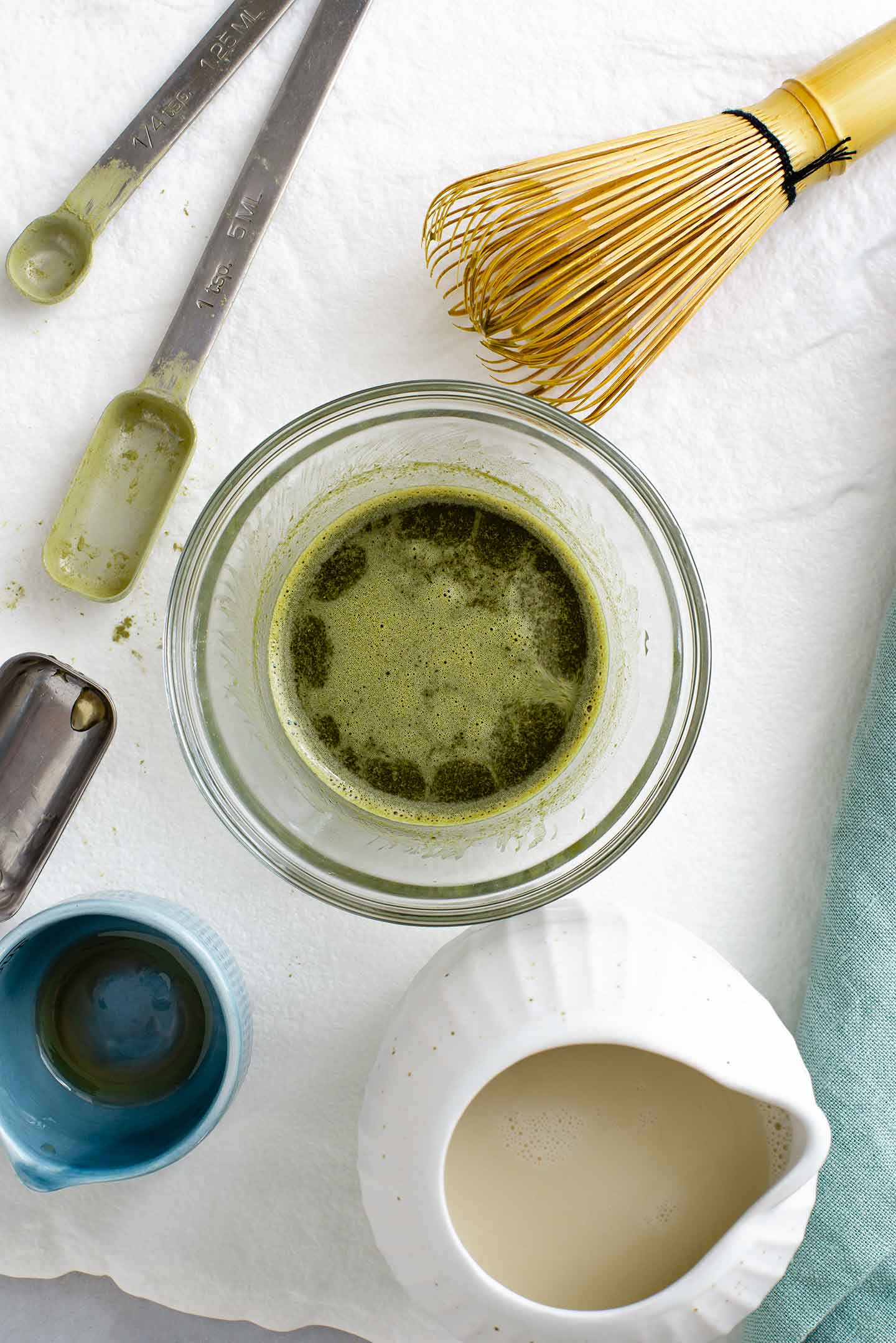 Top down view of the green tea mixture in a small glass bowl with a delicate foam on top. A just used, bamboo whisk rests next to it on a white tray.