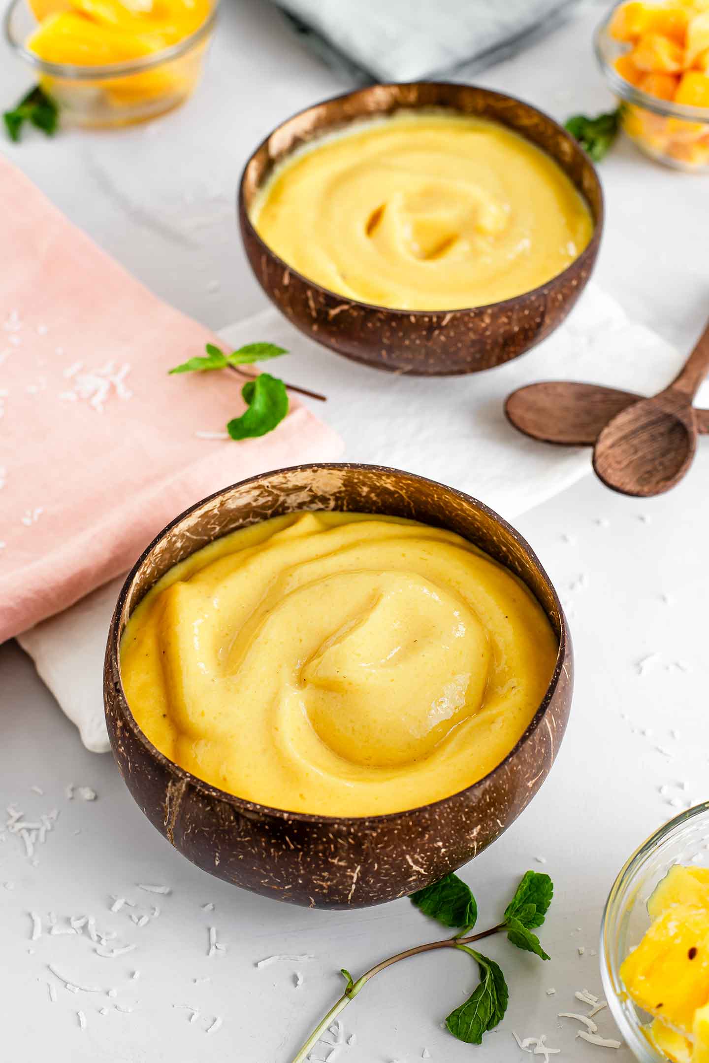 Top down view of two coconut bowls filled with bright yellow tropical jackfruit smoothies. Sprigs of mint, bamboo spoons and small bowls of mango, pineapple, and jackfruit surround the smoothies.