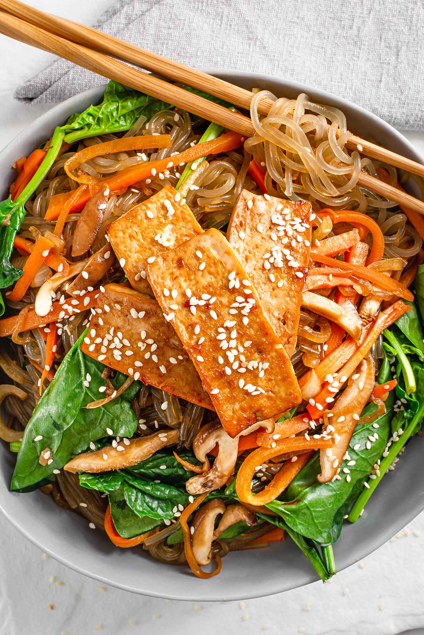 Top down view of simple vegan japchae in a bowl. Glass noodles are wrapped around chopsticks, carrots, onion, mushroom, and spinach mix with the noodles. Tofu and toasted sesame seeds top it off.