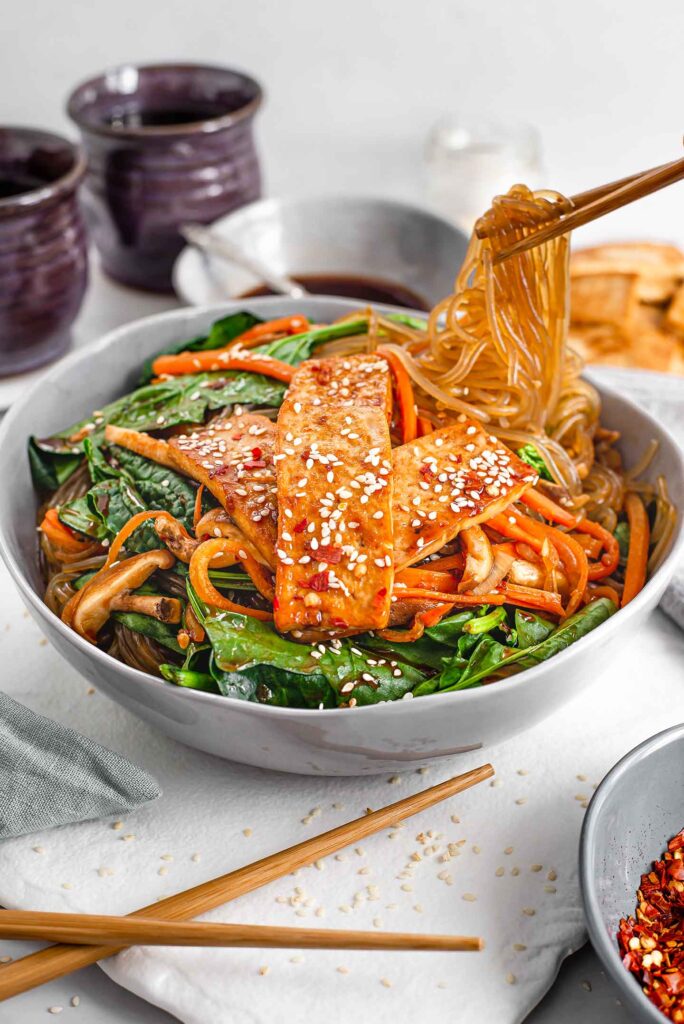 Side view of sweet potato noodles being lifted from a bowl of Japchae with chopsticks. The Japchae has spinach, carrot, onion, mushrooms, and tofu.
