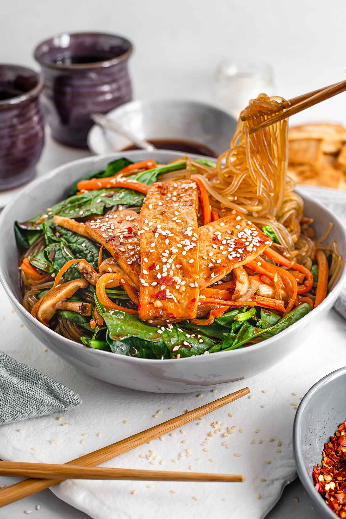 Side view of sweet potato noodles being lifted from a bowl of vegan Japchae with chopsticks. The Japchae has spinach, carrot, onion, mushrooms, and tofu.