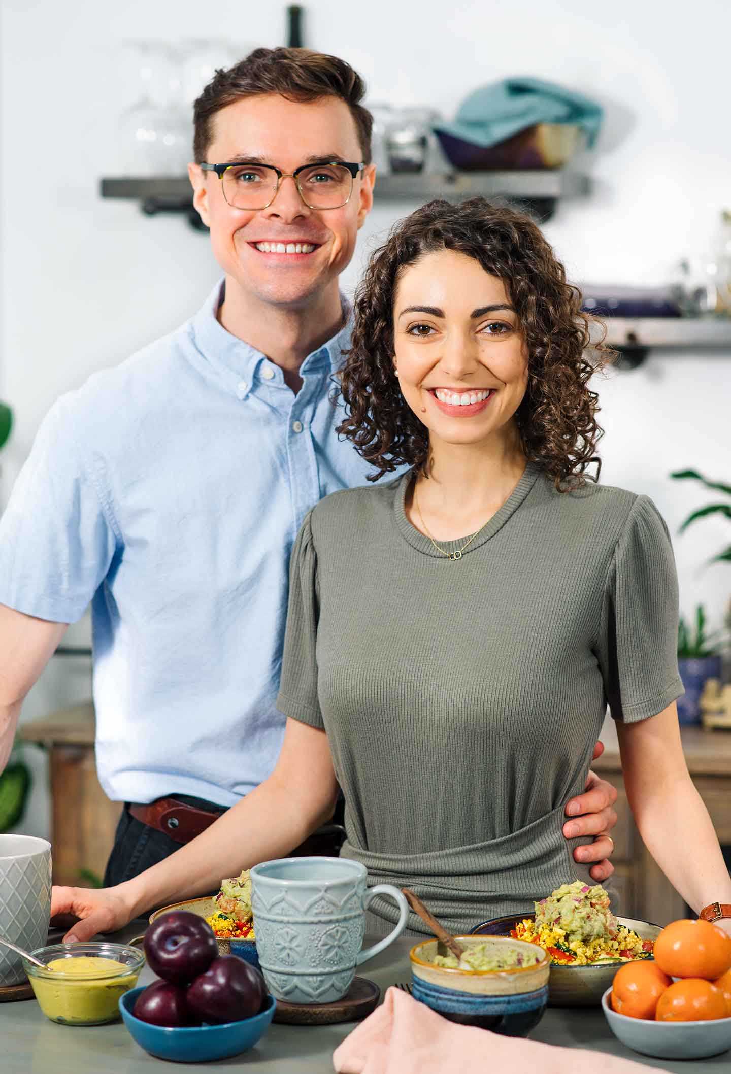 Bryan and Kathryn standing in their kitchen with tofu scramble, fruit, guacamole and coffee mugs sitting in front of them on a table.