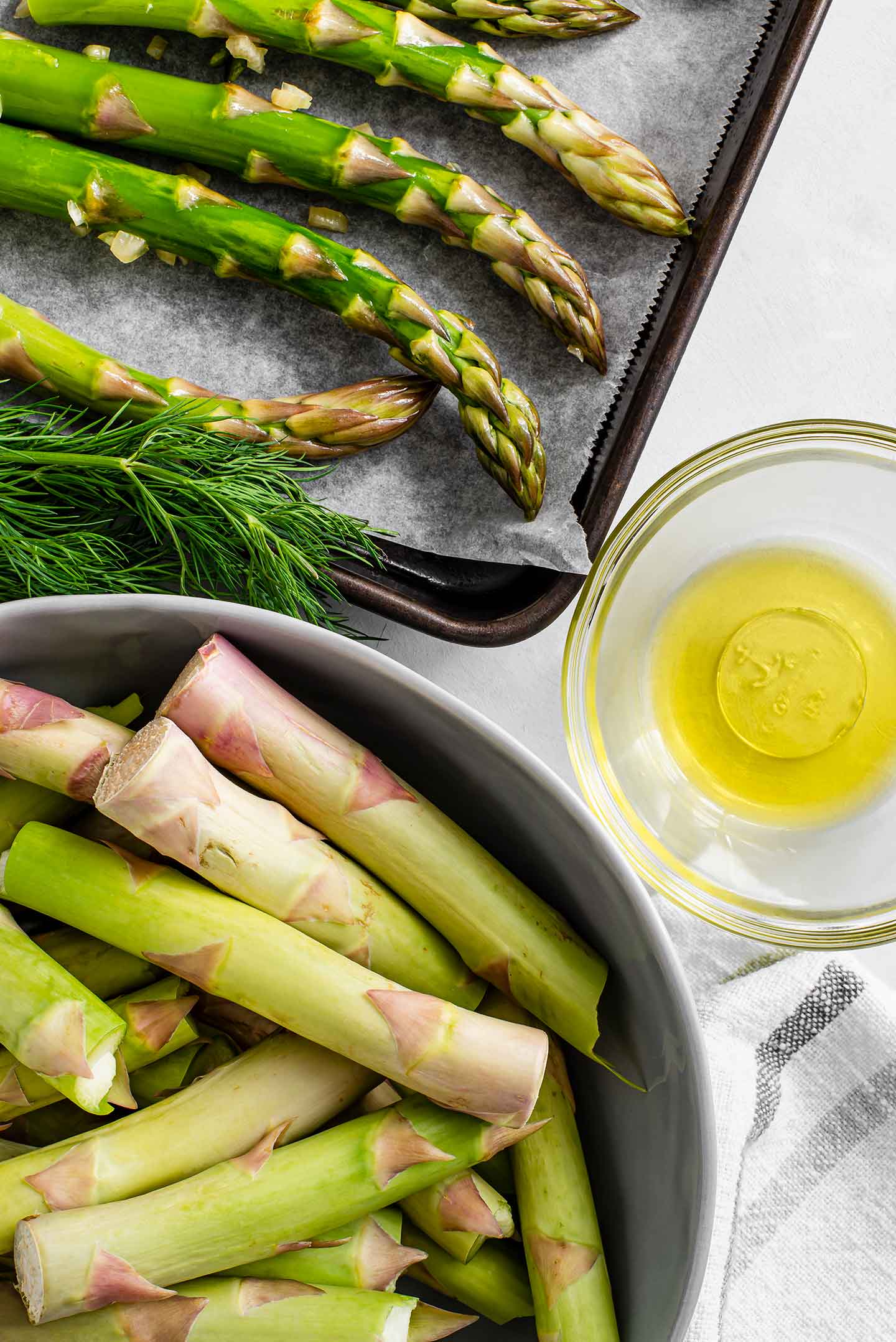 Top down view of asparagus spears on a lined baking tray with the tough ends in a separate bowl. A small dish of olive oil stands beside the baking tray.