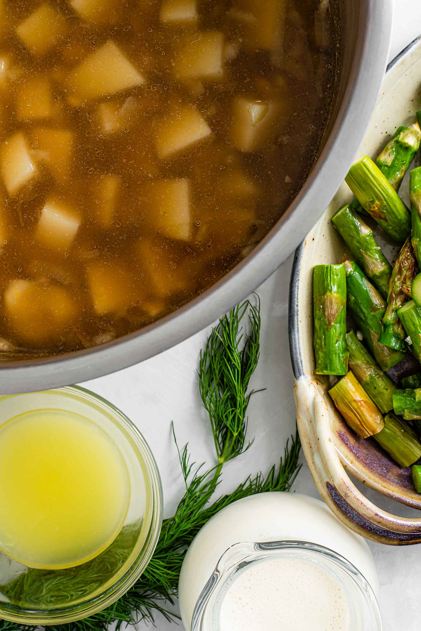 Top down view of a large pot with potatoes, beans, and veggie broth. Next to the pot is sliced roasted asparagus, lemon juice, plant milk, and fresh dill.