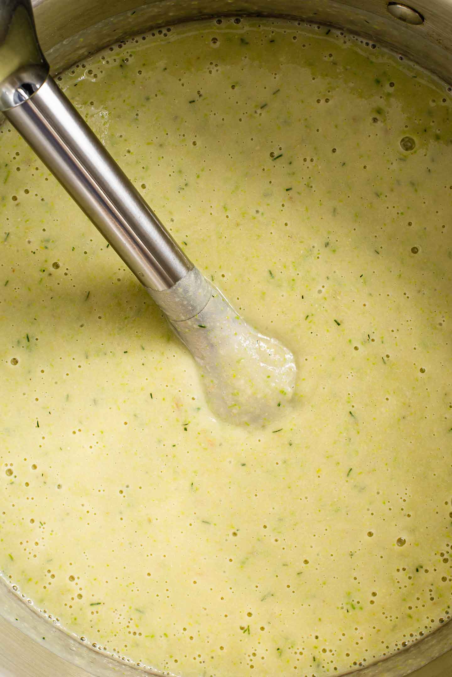 Top down view of an immersion blender in smooth, creamy soup. The soup is still speckled with bits of green from the fresh dill and asparagus.