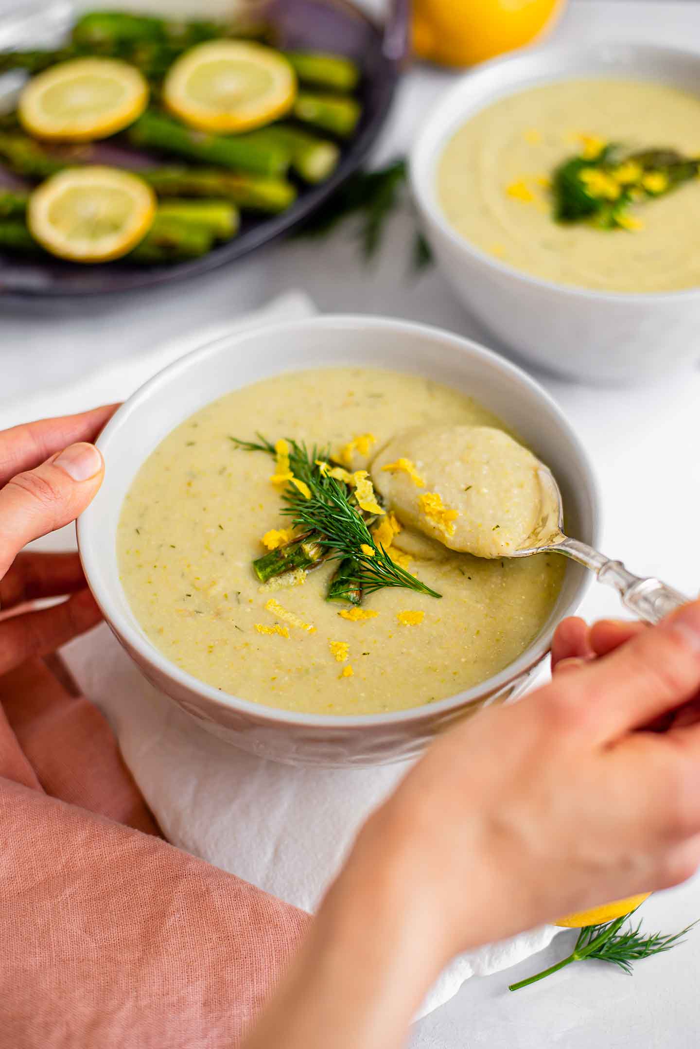 Side view of hand holding a spoon and scooping thick, creamy, lemon asparagus soup. The soup is garnished with asparagus, lemon zest, and dill. More asparagus spears and another bowl of soup are in the background.