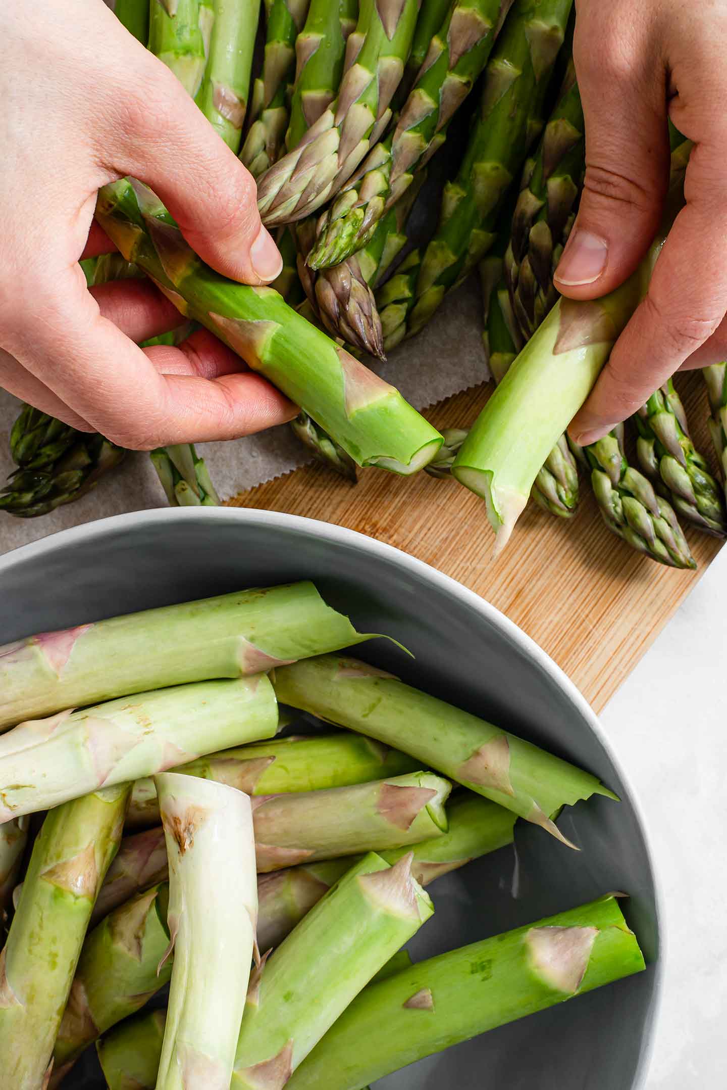 Top down view of two hands snapping a spear of asparagus. The pale, tough ends are collected in a small bowl and the fresh spears are laid on a wooden board.