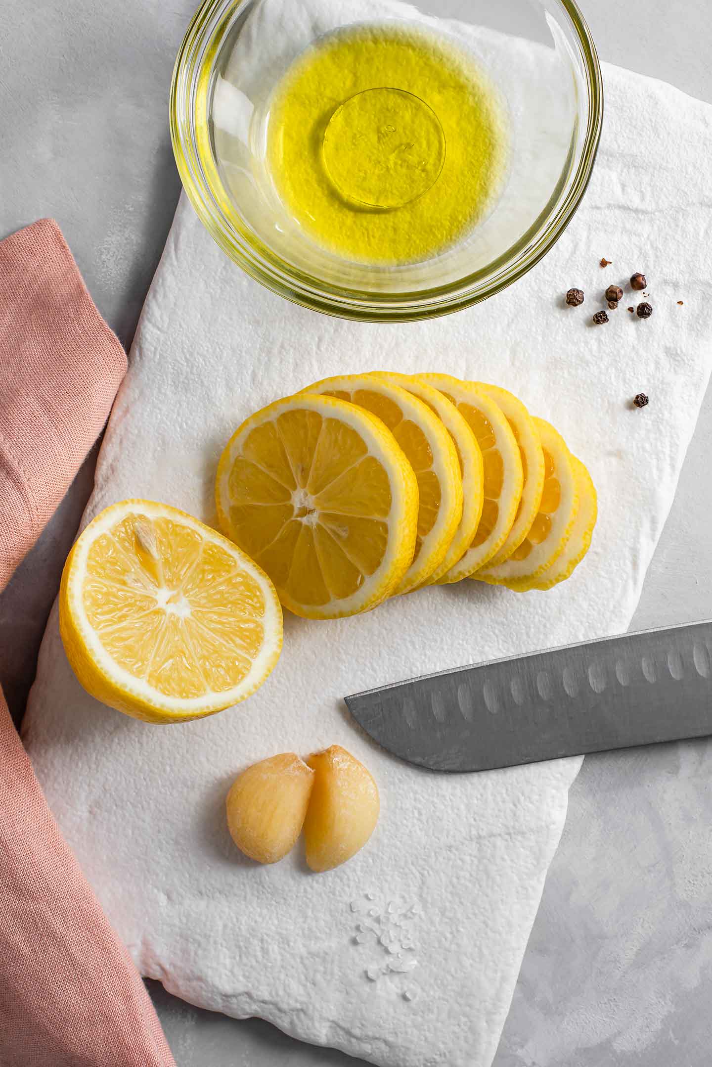 Top down view of thinly sliced lemon on a white tray with olive oil in a small jar, garlic cloves, and coarse salt and peppercorns.