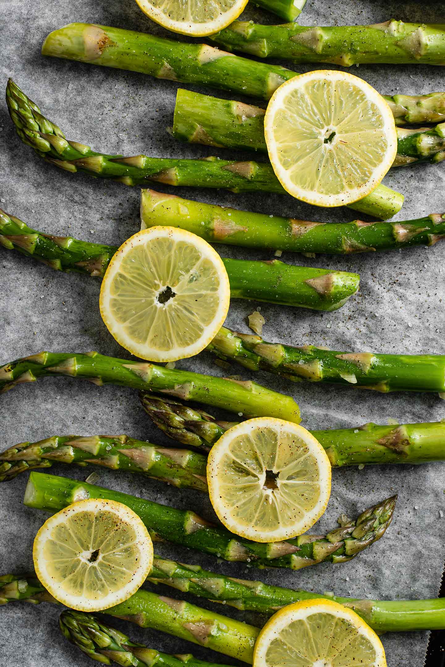 Top down view of oiled asparagus spears with garlic, salt, pepper, and thinly sliced lemon wedges laid in a single layer on a baking tray.
