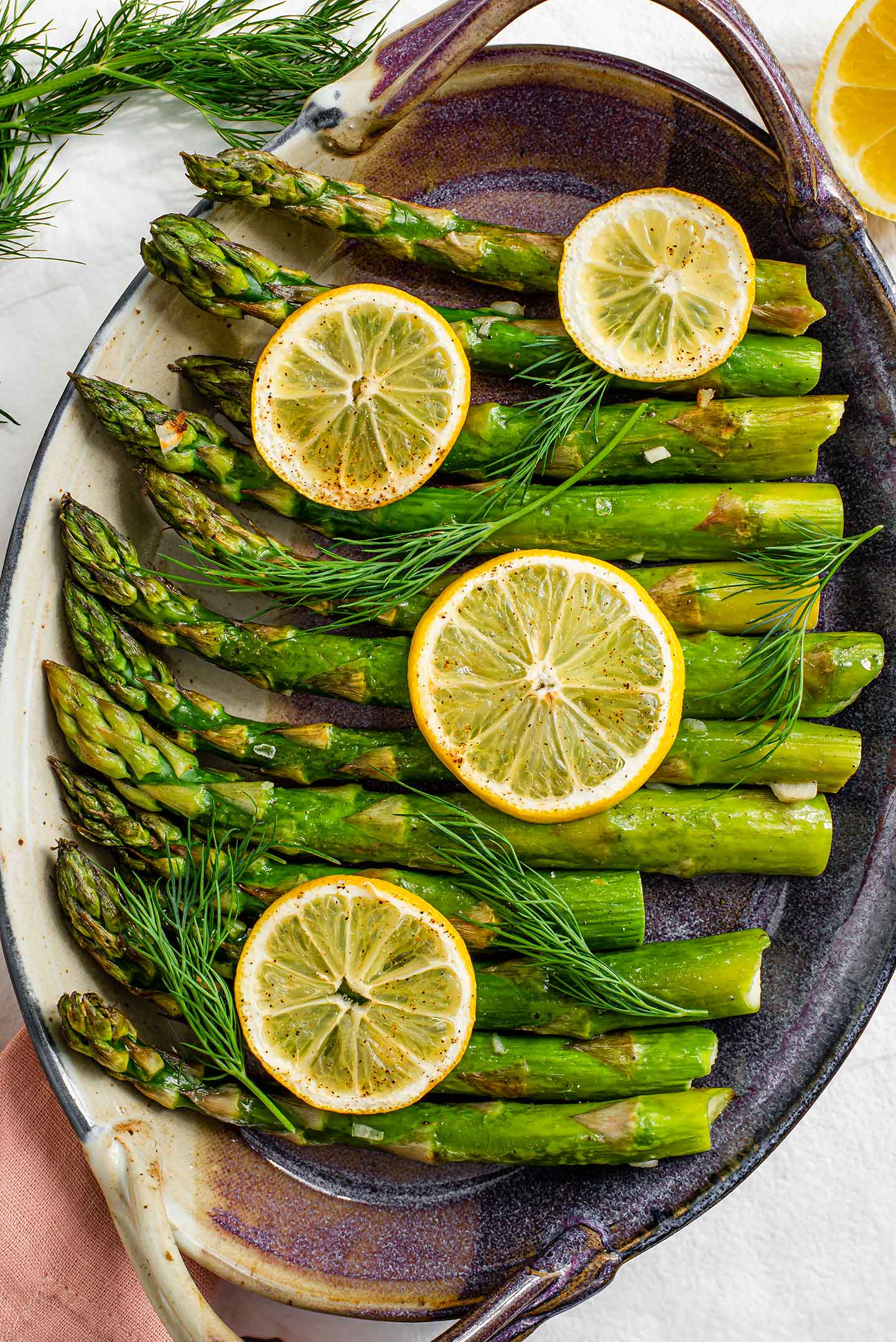 Top down view of lemon garlic roasted asparagus in a serving tray, garnished with lemon wedges and fresh dill. The spears are lightly oiled, charred, and speckled with minced garlic, salt, and pepper.