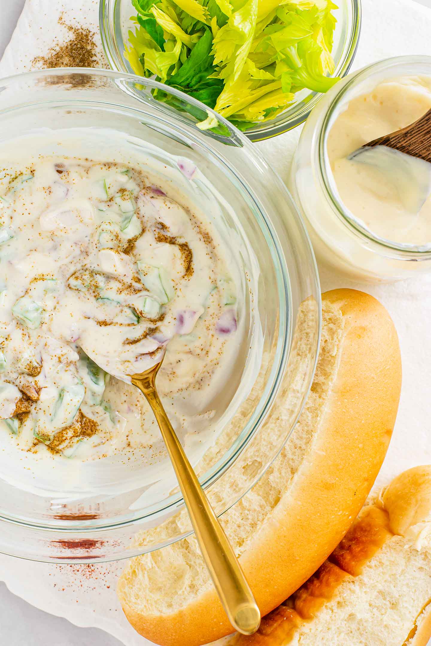 Top down view of oil-free mayonnaise mixed in a glass bowl with diced celery, red onion, lemon juice, and celery salt. New England style buns rests beside the bowl.