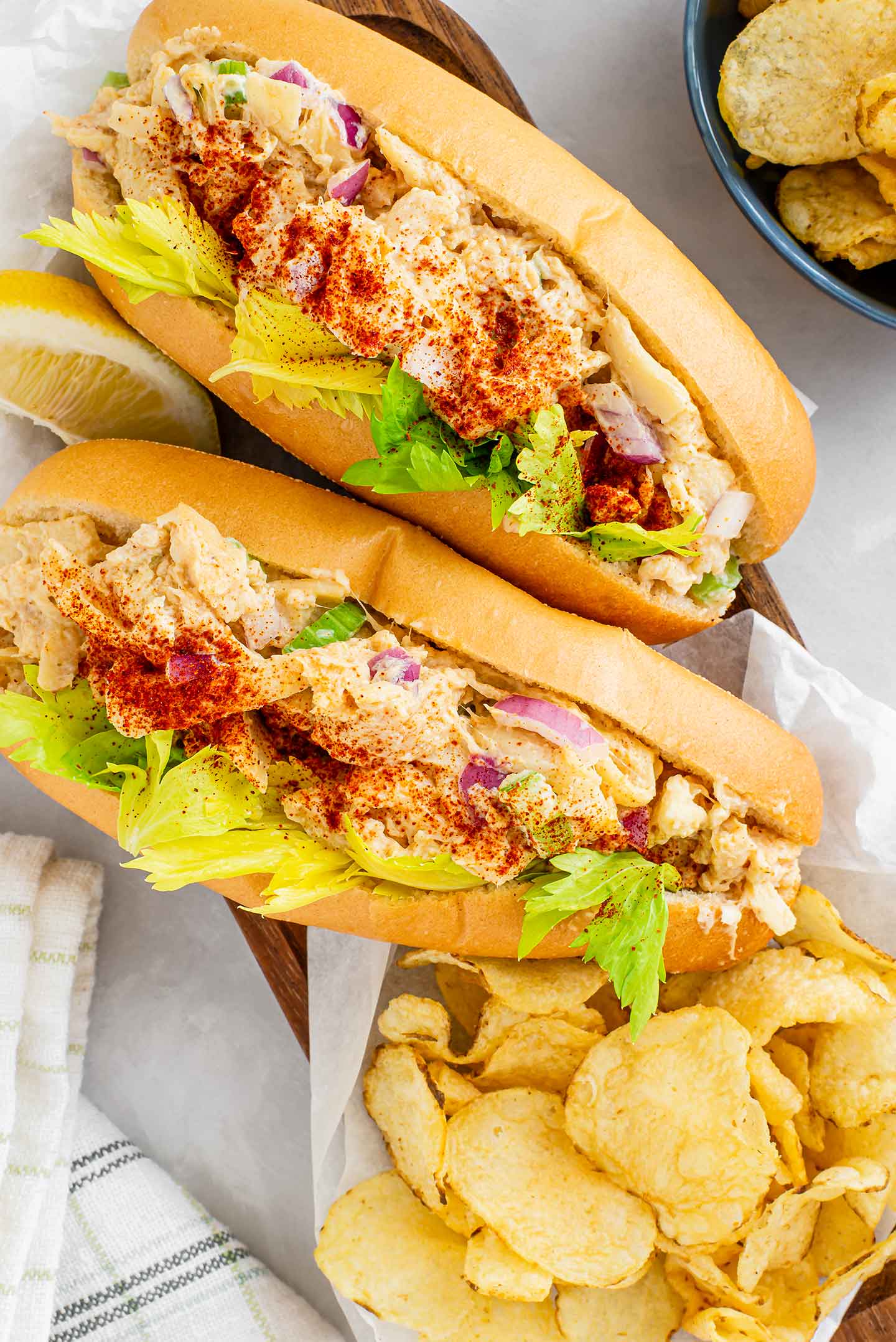 Top down view of two fish-less lobster rolls bursting with hearts of palm, sprinkled with paprika, garnished with celery leaves and served with a side of kettle chips.