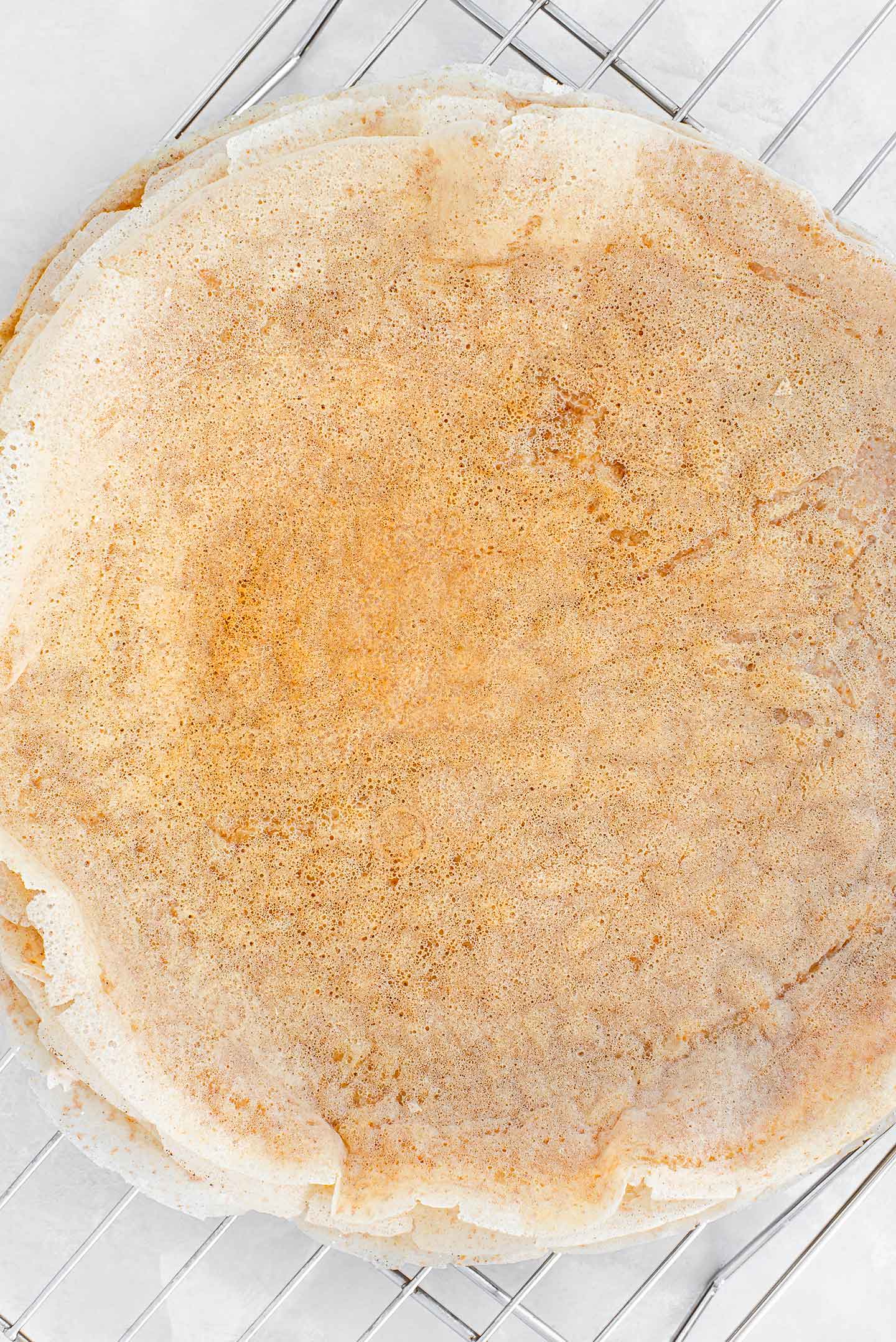 Top down view of a stack of cooked crepes cooling on a wire rack. The edges are crispy and the crepe is golden.