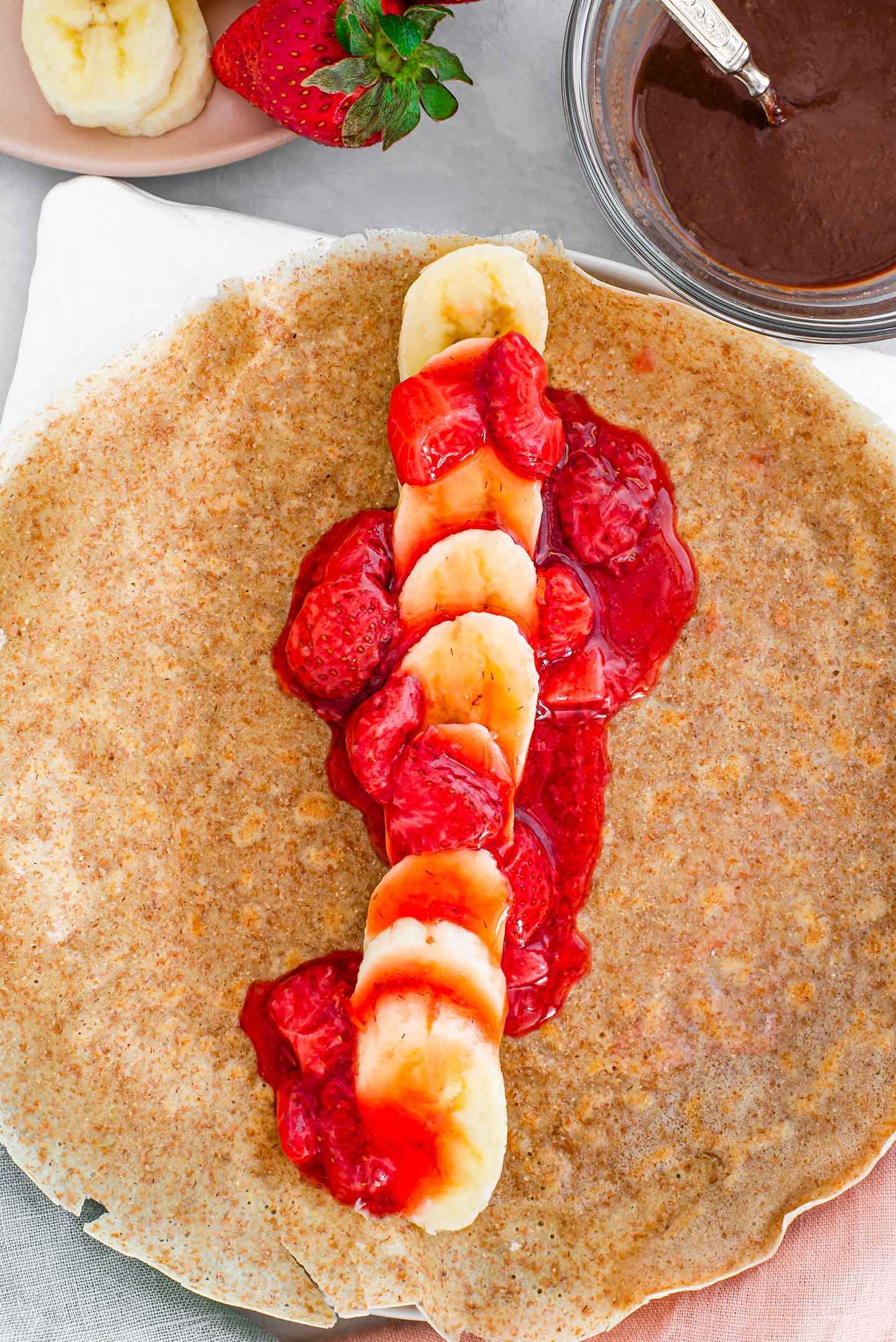 Top down view of sliced banana in a single layer in the middle of a crepe with strawberry sauce drizzled over before being rolled up.