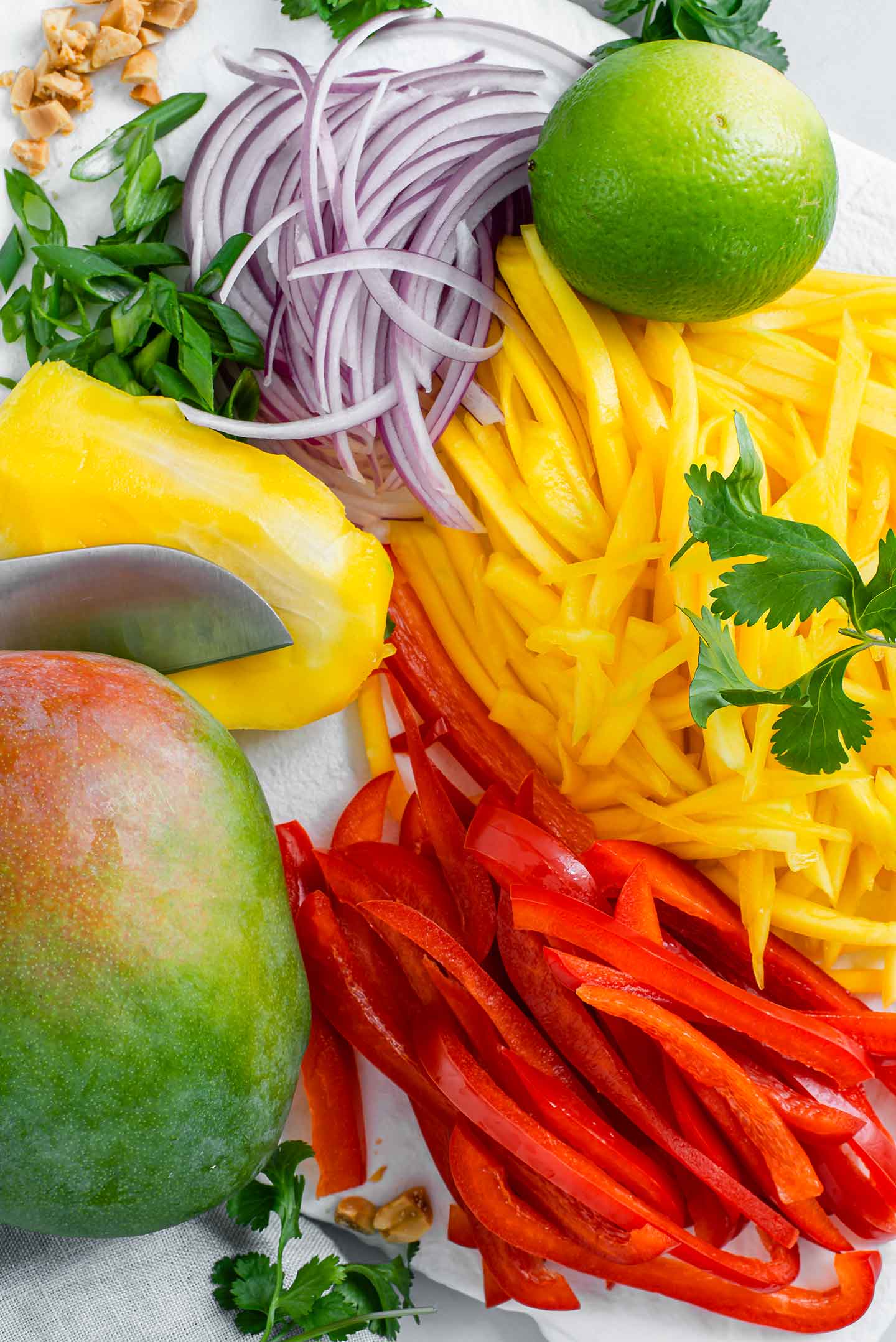 Top down view of a whole ripe mango, thinly sliced mango, sliced red pepper, sliced red onion, sliced green onion, a lime and cilantro leaves.