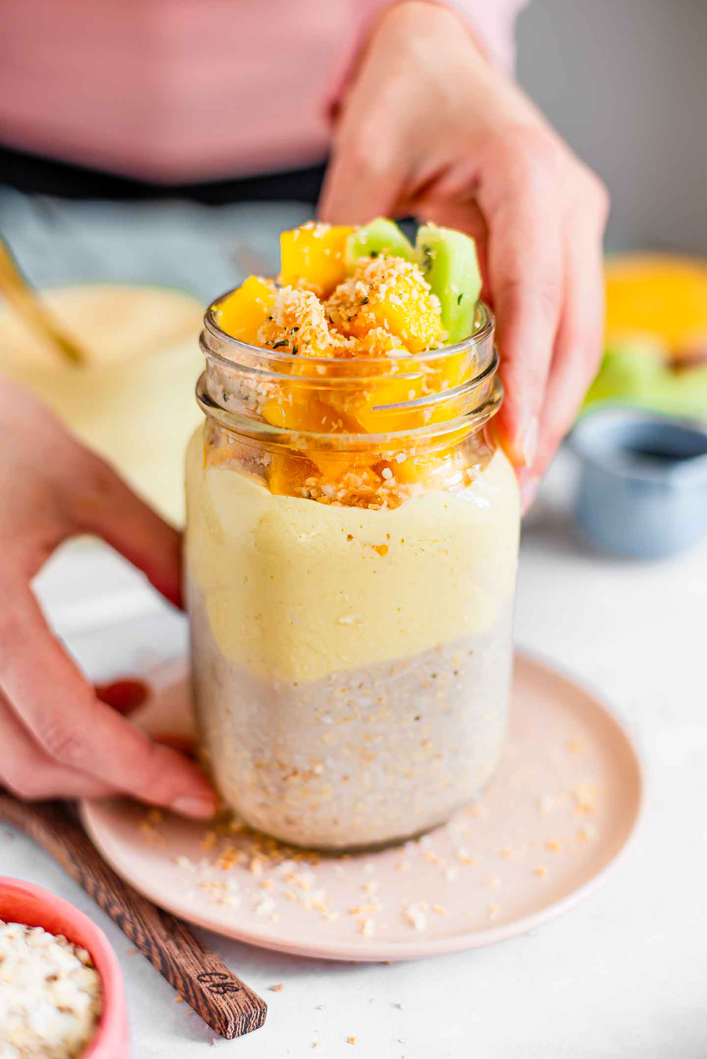 Side view of hands placing a colourful jar of mango yogurt overnight oats on a small plate. The oats are visible in the bottom half. Then a layer of mango yogurt is visible. And fresh mango, toasted coconut, hemp seeds, and some slices of kiwi sit on top.
