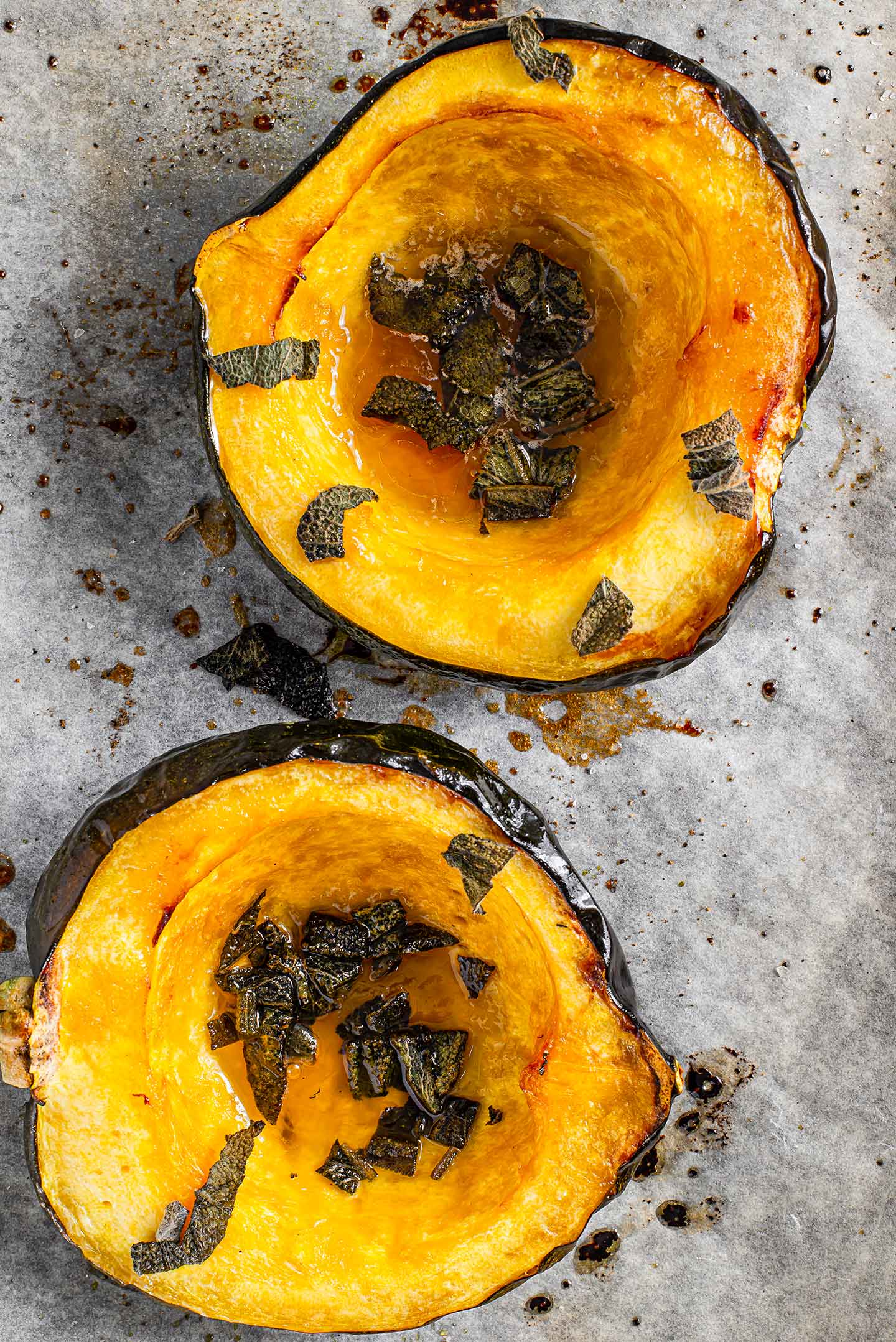 Top down view of maple roasted acorn squash halves on a parchment lined baking tray. The halves are caramelized, butter and maple syrup are pooled in the hollow centre, and sage leaves are scattered over the flesh.