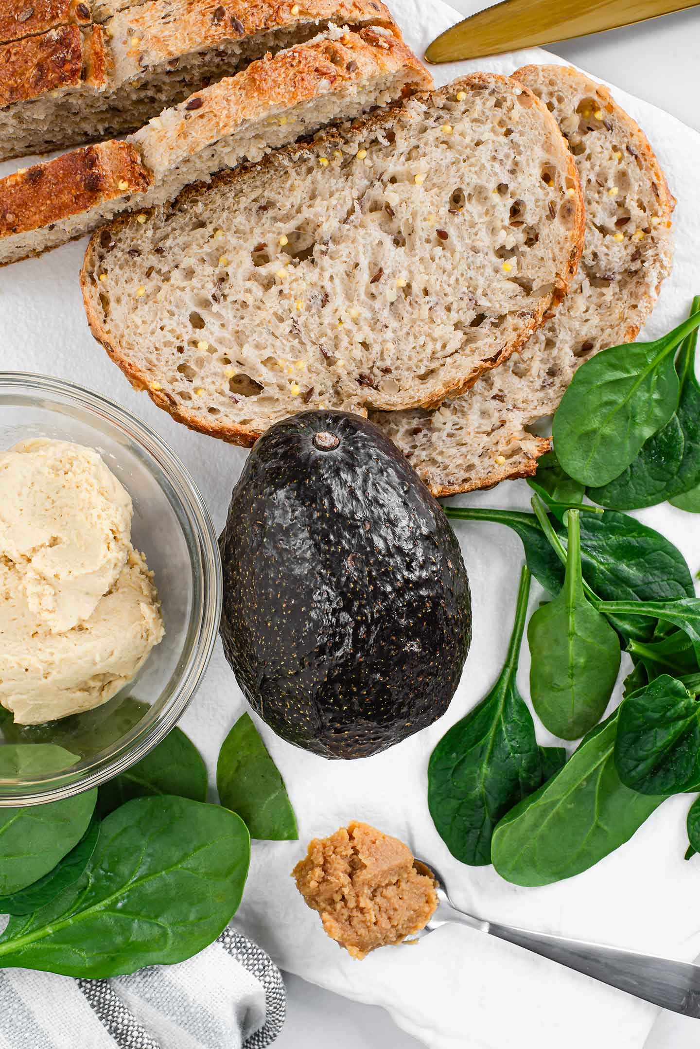 Top down view of an avocado surrounded by spinach leaves, miso paste on a spoon, a small bowl of hummus, and slices of seeded bread.