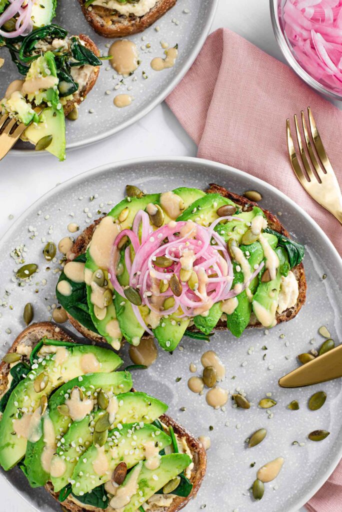 Top down view of two slices of miso avocado toast on a plate. Topped with seeds and pickled red onion. A fork and napkin lay beside. Another plate of toast visible in the corner.