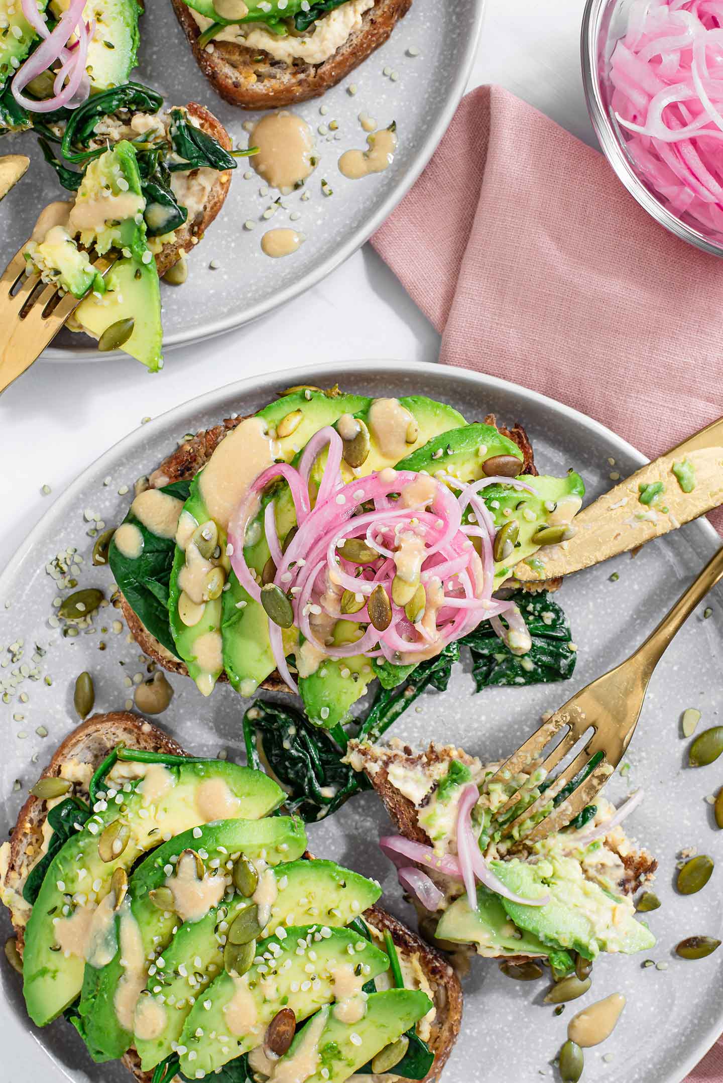Top down view of two slices of miso avocado toast on a plate with a fork and knife digging in. The toast is topped with pickled red onion, seeds, and drizzled with miso glaze. Two more slices are on another plate.