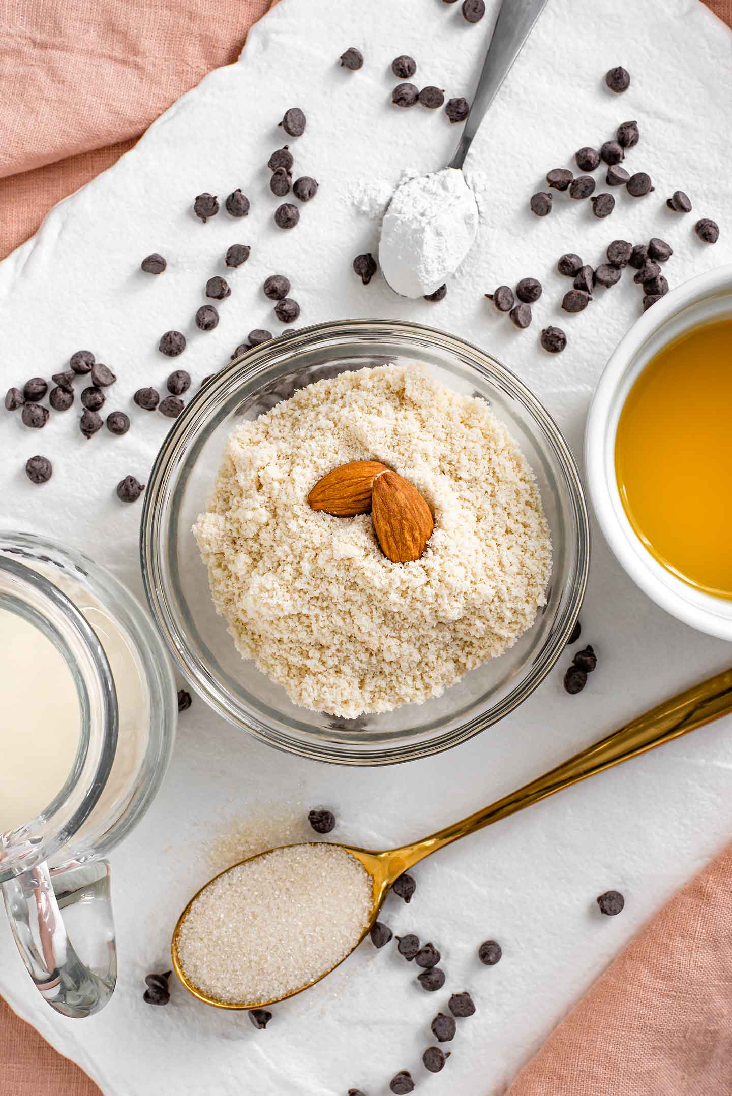 Top down view of almond flour in a small bowl with two almonds placed on top. Melted butter, milk, sugar, baking powder, and chocolate chips are scattered around.