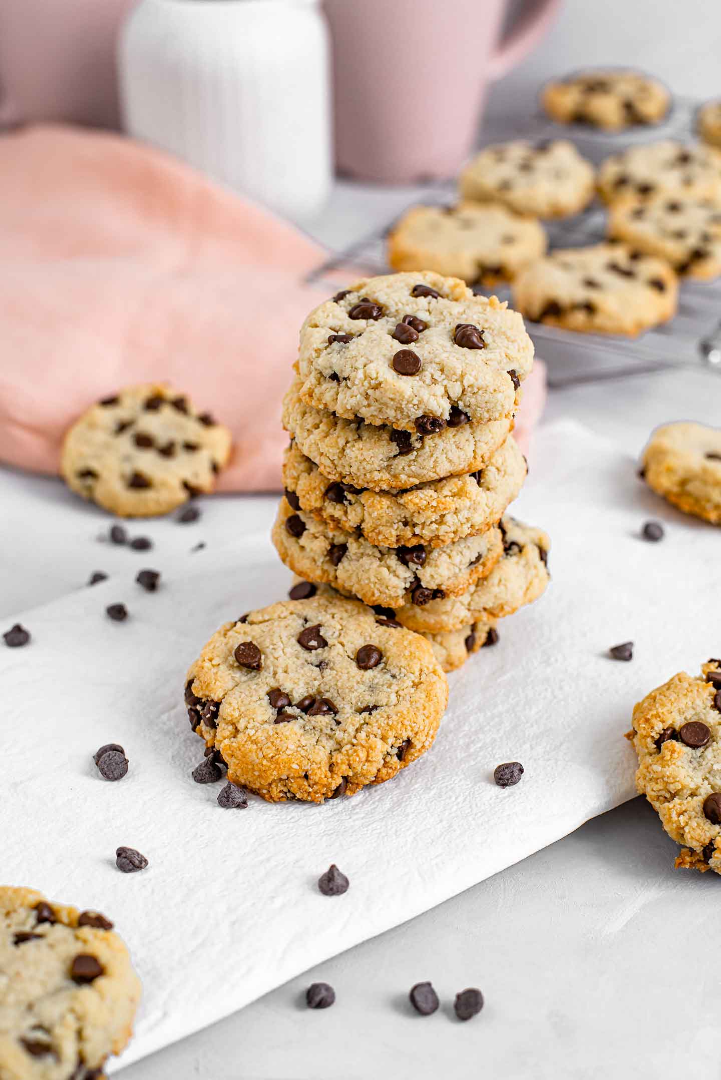 A tower of golden chocolate chip cookies on a white tray with chocolate chips and other cookies scattered around.