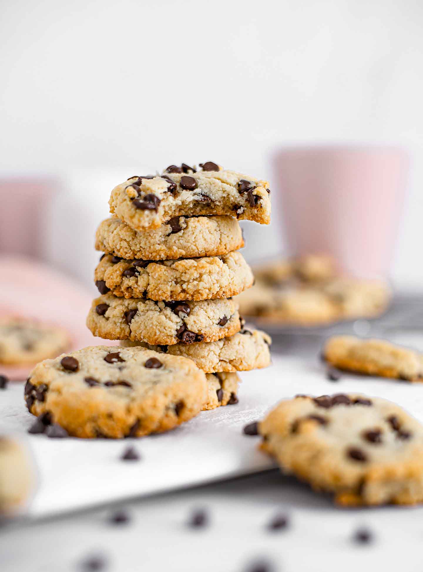 Close-up side view of a stack of cookies. The top cookie has a bite taken out of it. Mugs of coffee, a creamer, and more cookies are in the background.