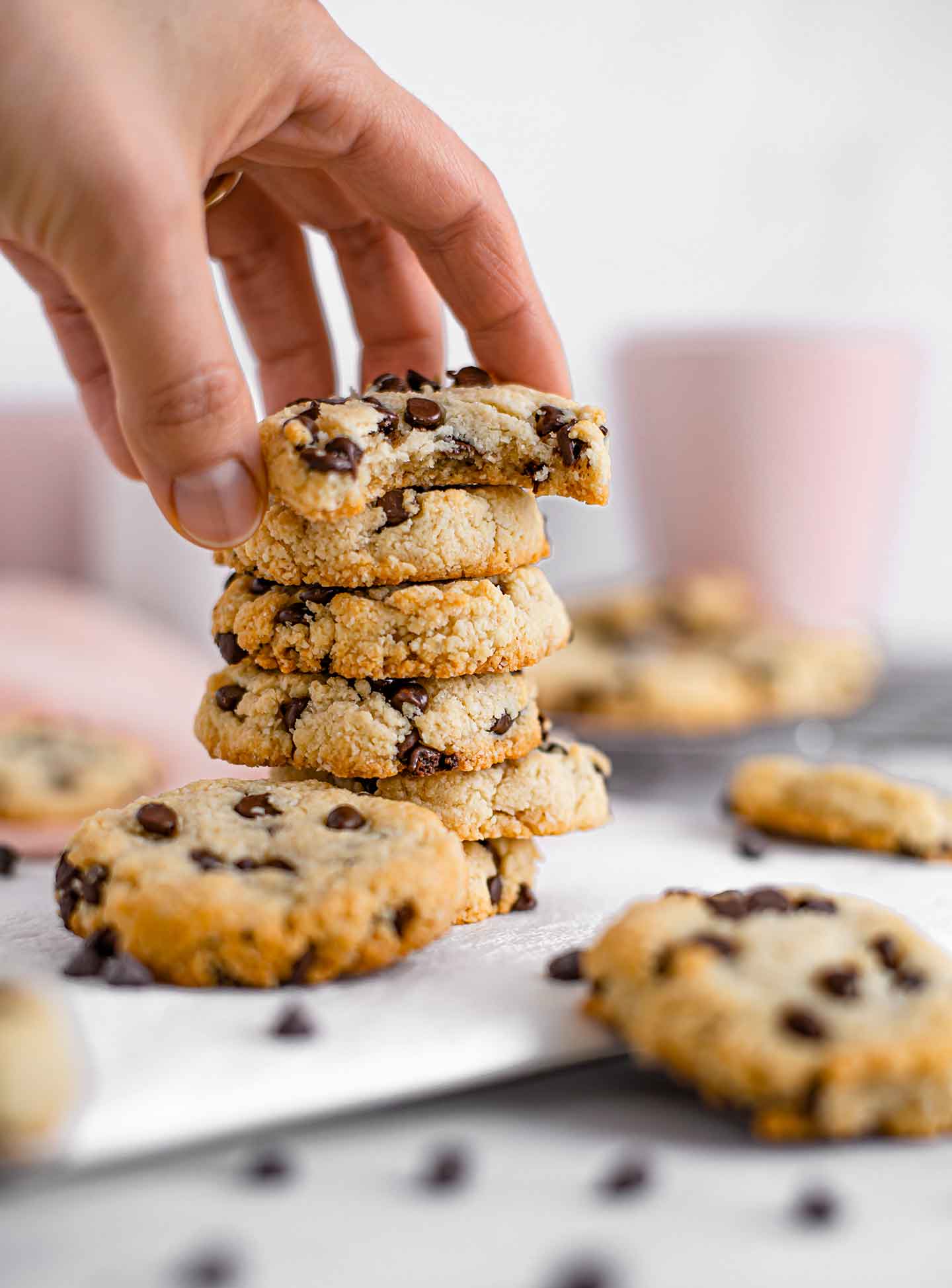 A hand grabs the top cookie off a stack. The cookie has a bite taken out and is filled with chocolate chips. Pink mugs fill the background and more cookies are scattered around.