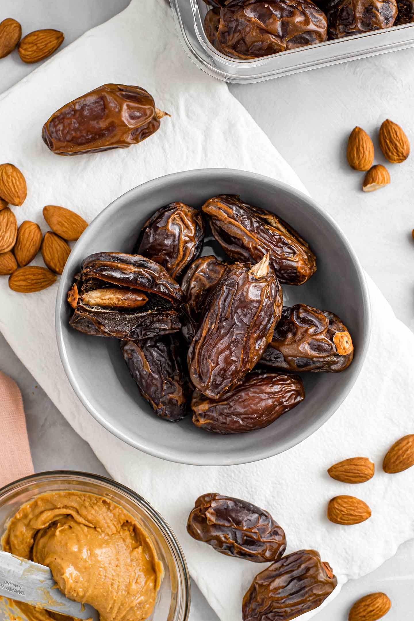 Top down view of dates filling a small bowl. One date is sliced open, exposing the put inside. Peanut butter and almonds surround the bowl of dates.