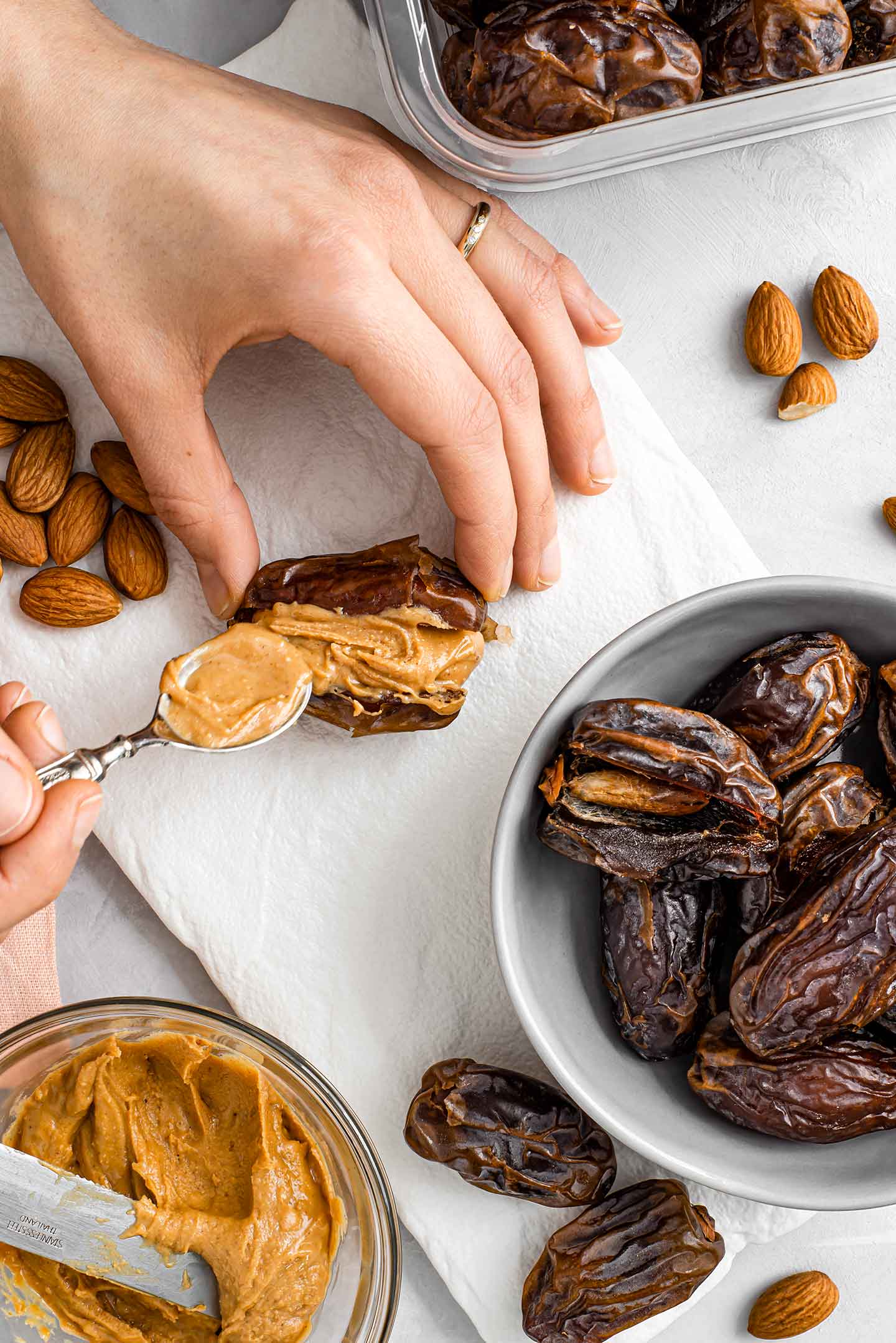 A hand spoons peanut butter into the centre of a medjool date. A bowl of other dates, loose almonds, and a bowl of peanut butter surround the hands.