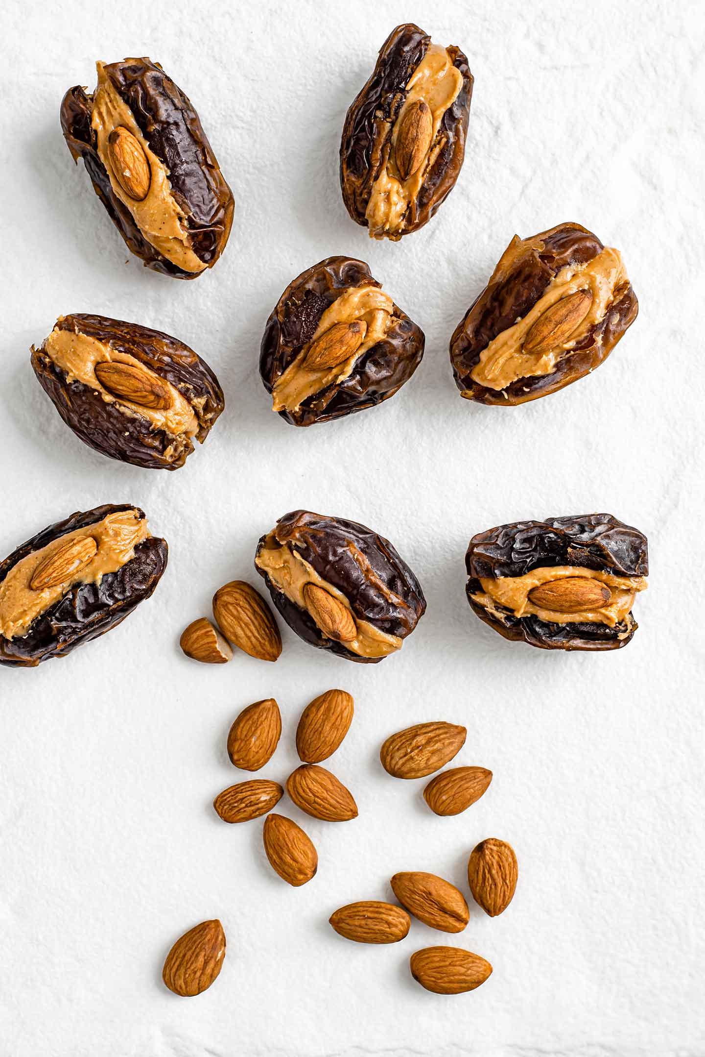 Top down view of eight medjool dates filled with peanut butter and raw almonds. Loose raw almonds cascade down a white tray.