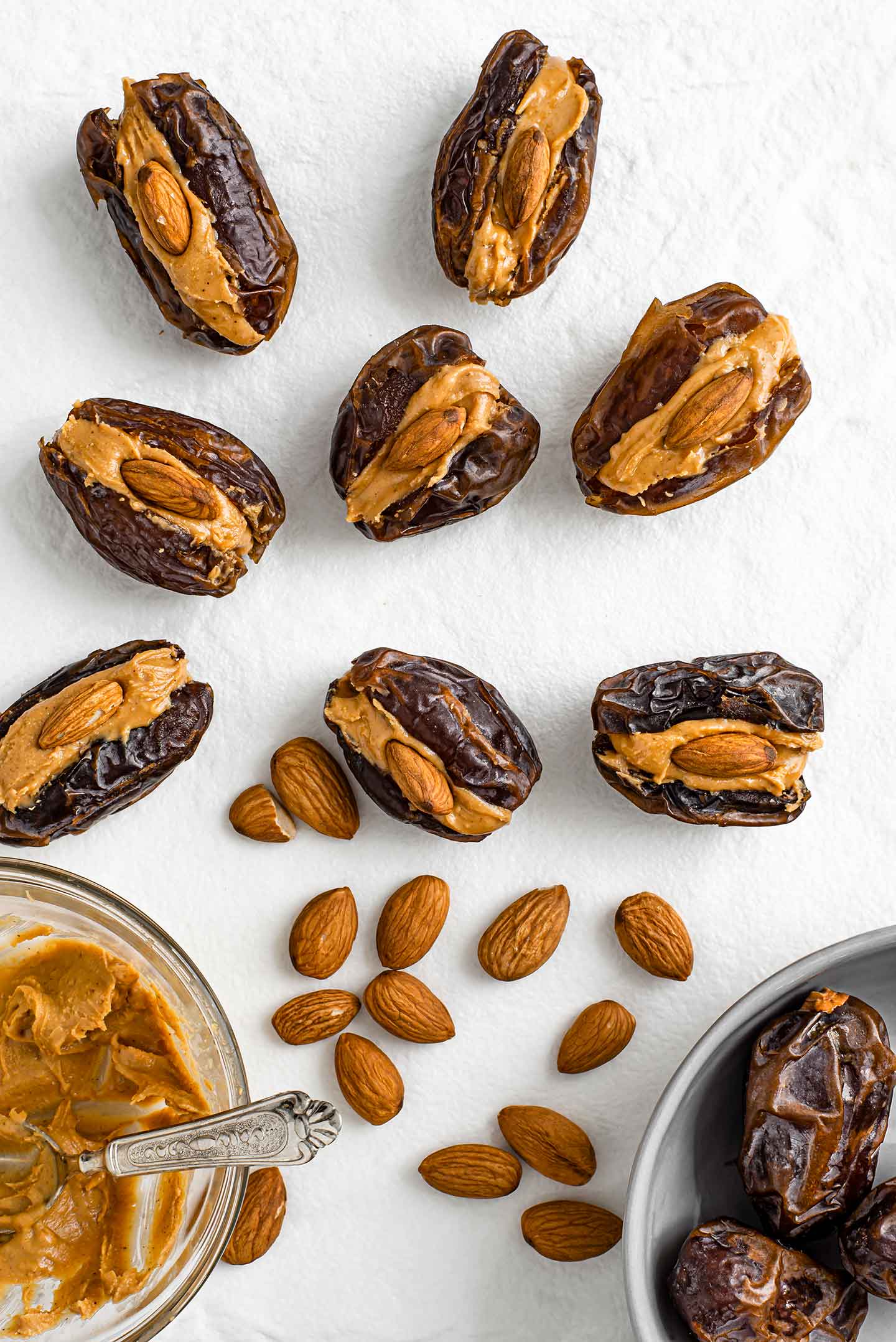 Peanut butter filled dates with almonds in the centre are scattered on a white tray. Loose almonds, a bowl of medjool dates, and a bowl of peanut butter are also pictured.