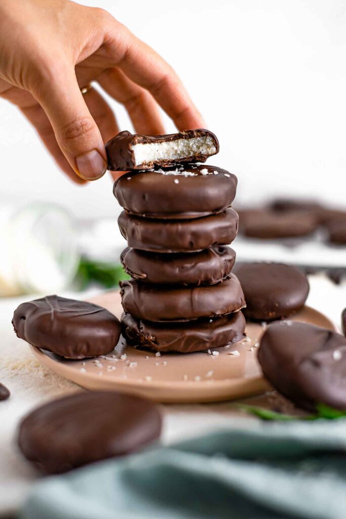 Side view of a stack of peppermint bounty patties. A bite is missing from the top patty showing the thick coconut centre. A hand rests the top patty on the stack.