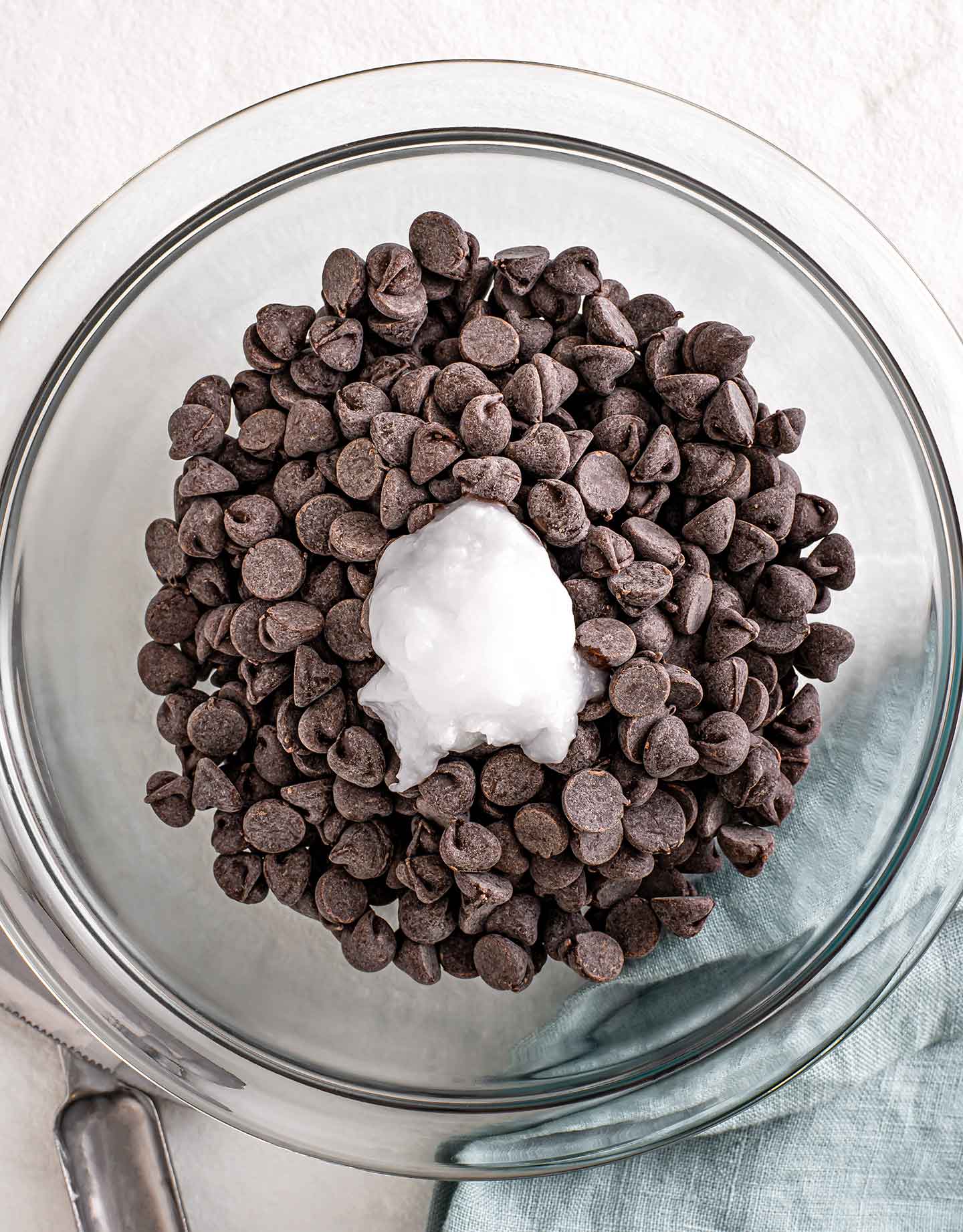Top down view of chocolate chips in a glass bowl with a scoop of coconut oil in the middle.