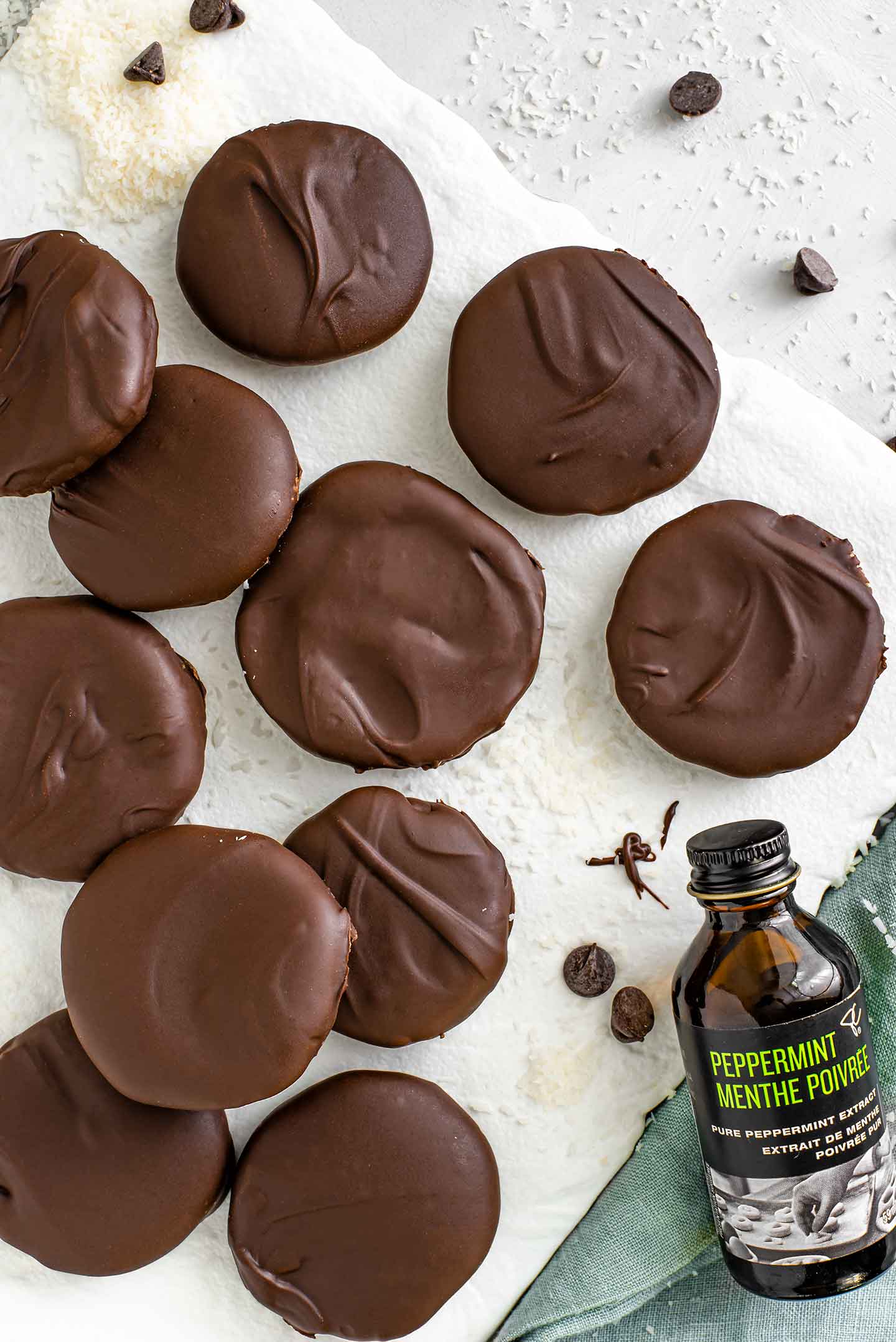 Top down view of coconut peppermint patties scattered across a white tray. Coconut flakes and chocolate chips are sprinkled around. A bottle of peppermint extract leans against the tray.