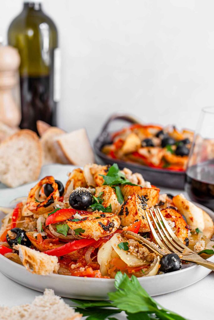 Side view of Portuguese Bacalhau on a plate with a fork resting on the side. Another plate of stew, a glass of red wine, a wine bottle, and slices of bread are in the background.