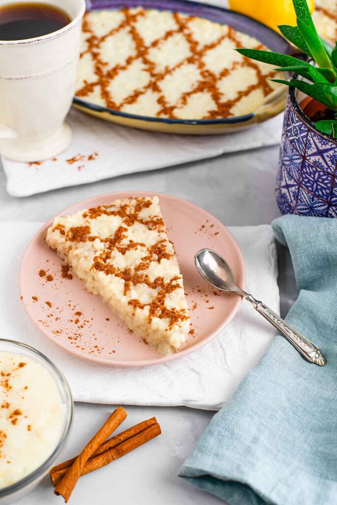 A pie shaped slice of Portuguese sweet rice sits on a small plate with a spoon. A full dish of decorated rice is in the background with a cup of coffee. Cinnamon sticks and a personal sized pudding dish are in the foreground.