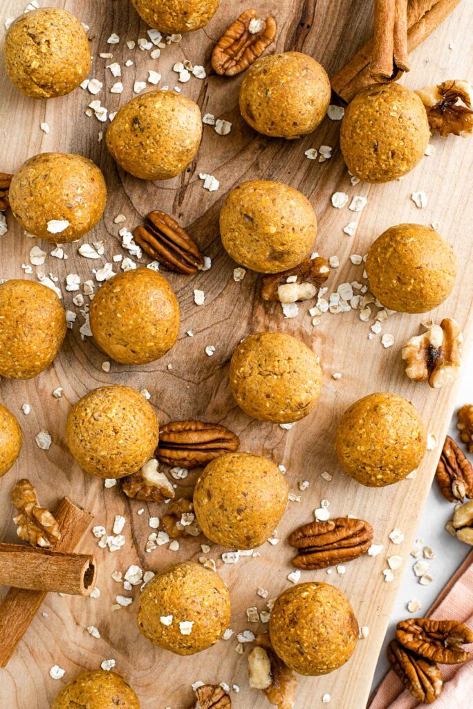 Top down view of pumpkin pie energy balls displayed on a wooden tray surrounded by oat flakes and pecans and cinnamon sticks.