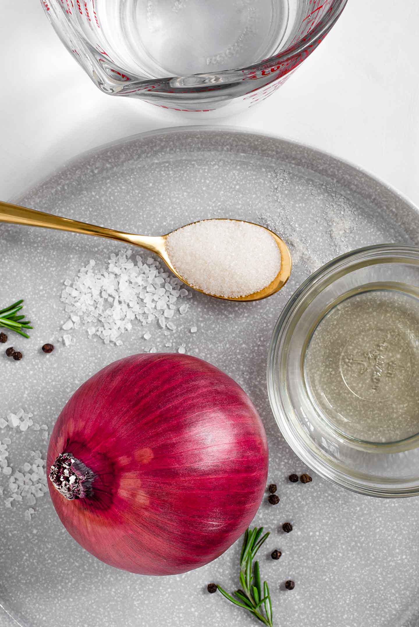 Top down view of a red onion on a large plate with a small bowl of vinegar, a spoon of sugar, a measuring cup of water, and sprigs of rosemary and peppercorns scattered around.