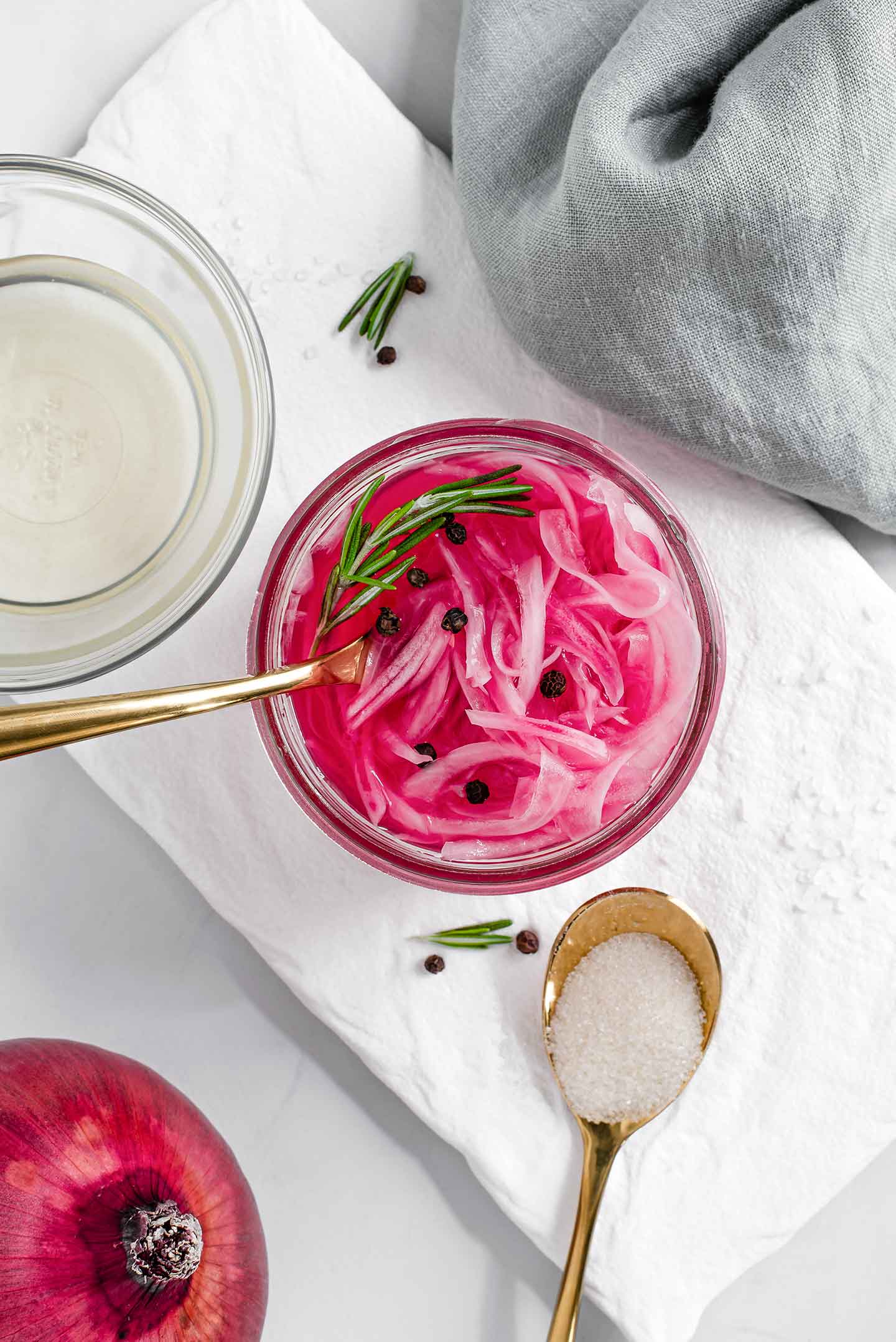 Top down view of quick pickled red onions in a glass jar with vinegar, sugar, salt, and a whole red onion on a white tray.
