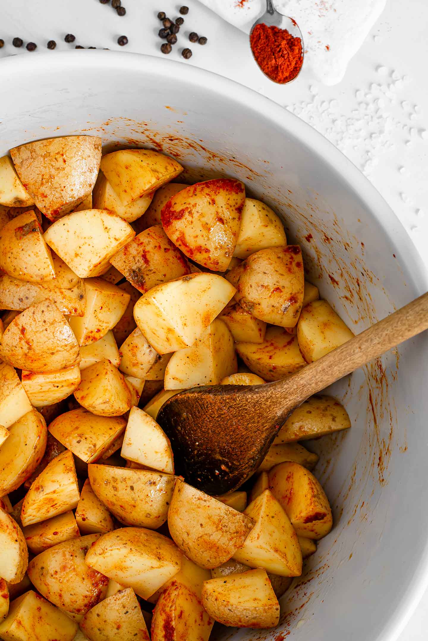 Top down view of raw diced potatoes in a large bowl, sprinkled with olive oil, smoked paprika, sat, pepper, and garlic. A wooden spoon rests in the bowl.