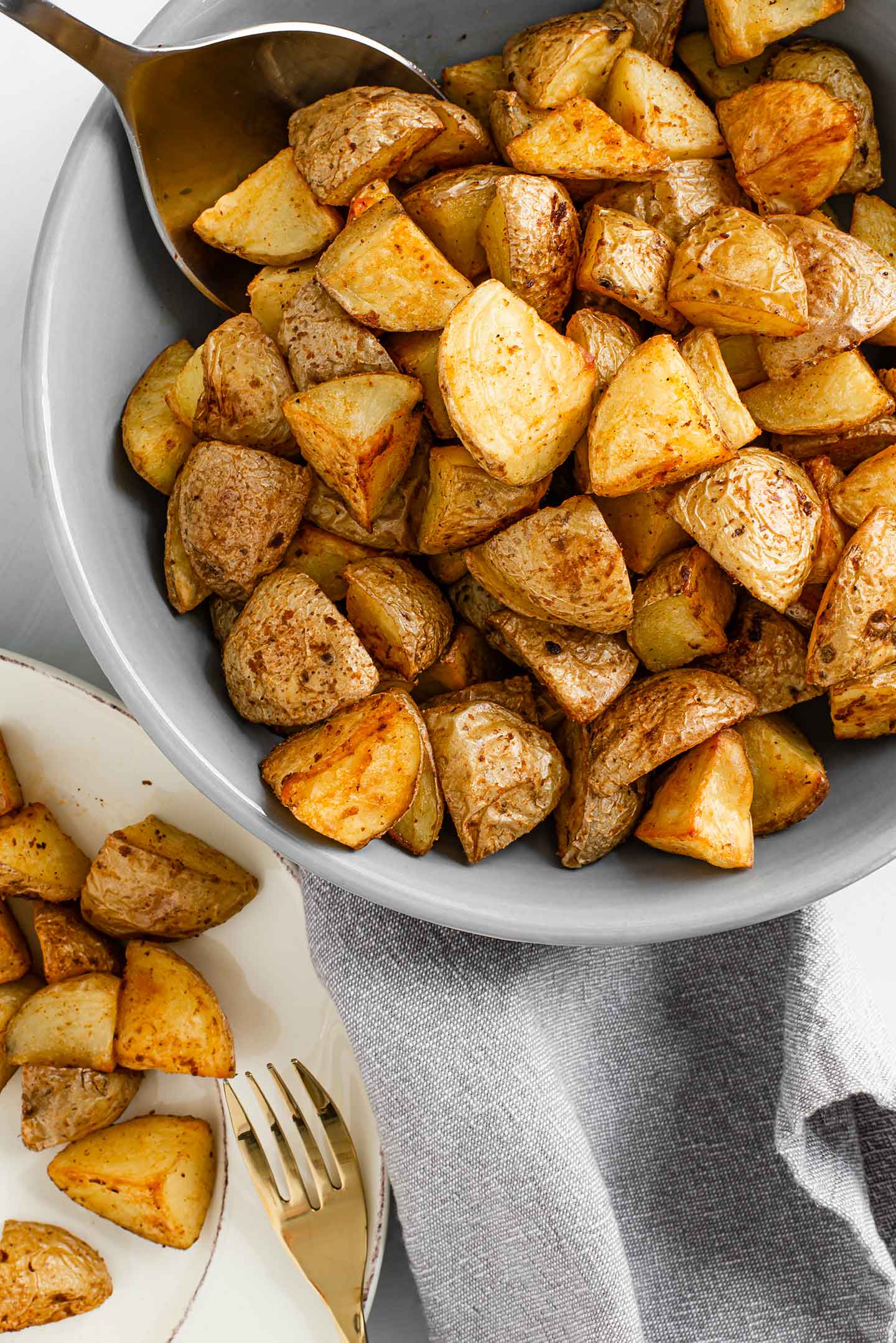 Top down view of crispy roasted potatoes in a bowl with a serving spoon. Just visible in the corner of the image is a plate with potatoes and a gold fork.