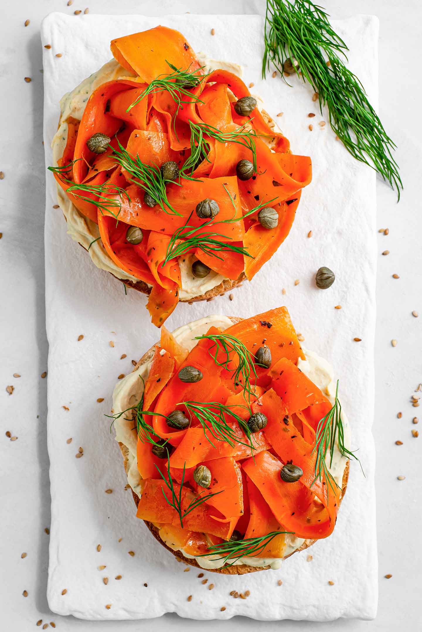 Top down view of two open-faced smoky lox breakfast bagels. The vibrant carrot lox is curled like smoked salmon and glistening from the marinade. Capers and dill garnish the top. The two halves are presented on a white tray.