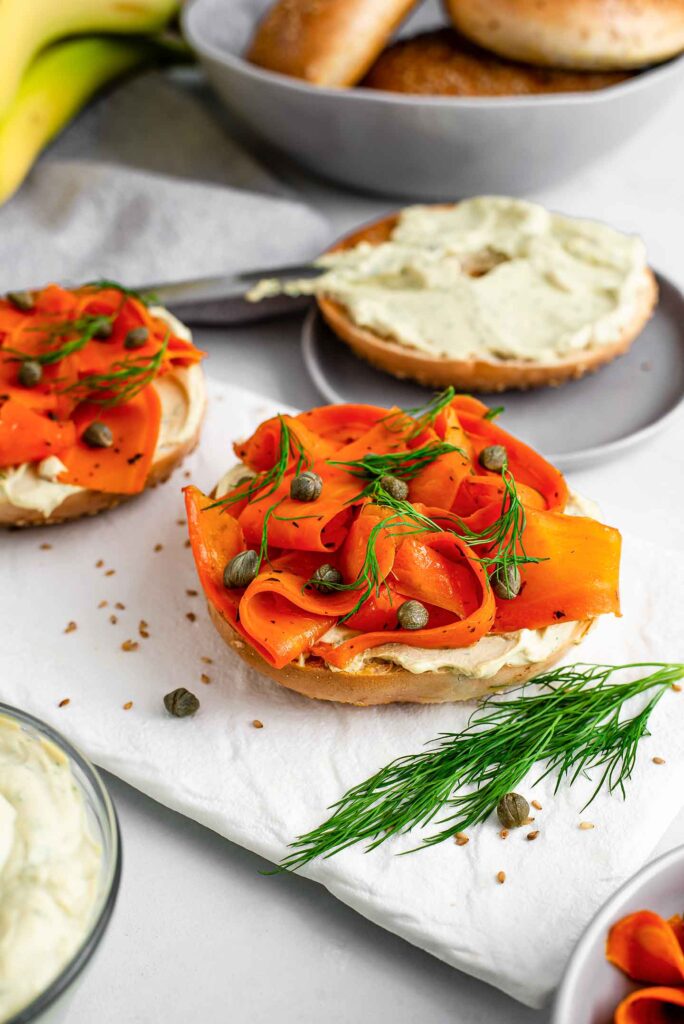 Side view of open-faced smoky lox breakfast bagels in the foreground. A half a bagel is being spread with creamy dill spread in the background. More bagels and some fruit surround.