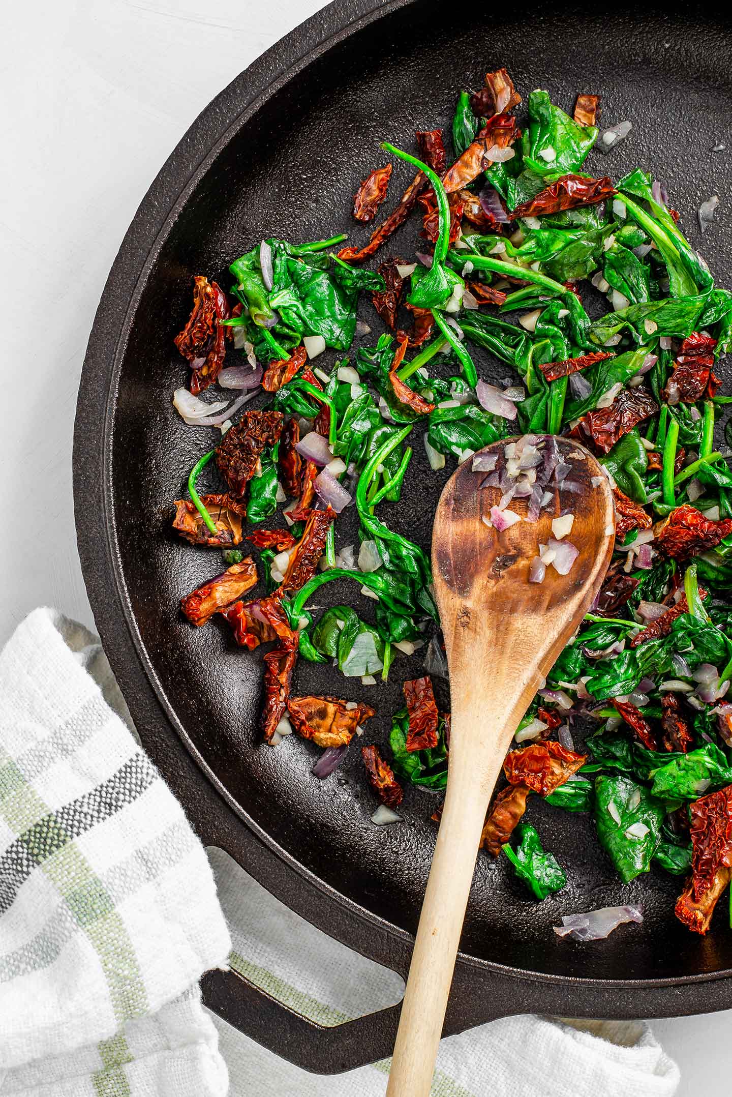 Top down view of sun-dried tomatoes, wilted spinach, cooked onion, and garlic in a cast iron skillet with a wooden spoon.
